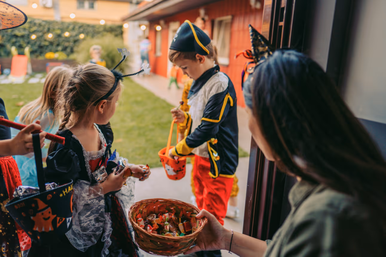 Children dressed as pirates and witches trick-or-treat at a brightly lit house as an adult offers candy from a basket.