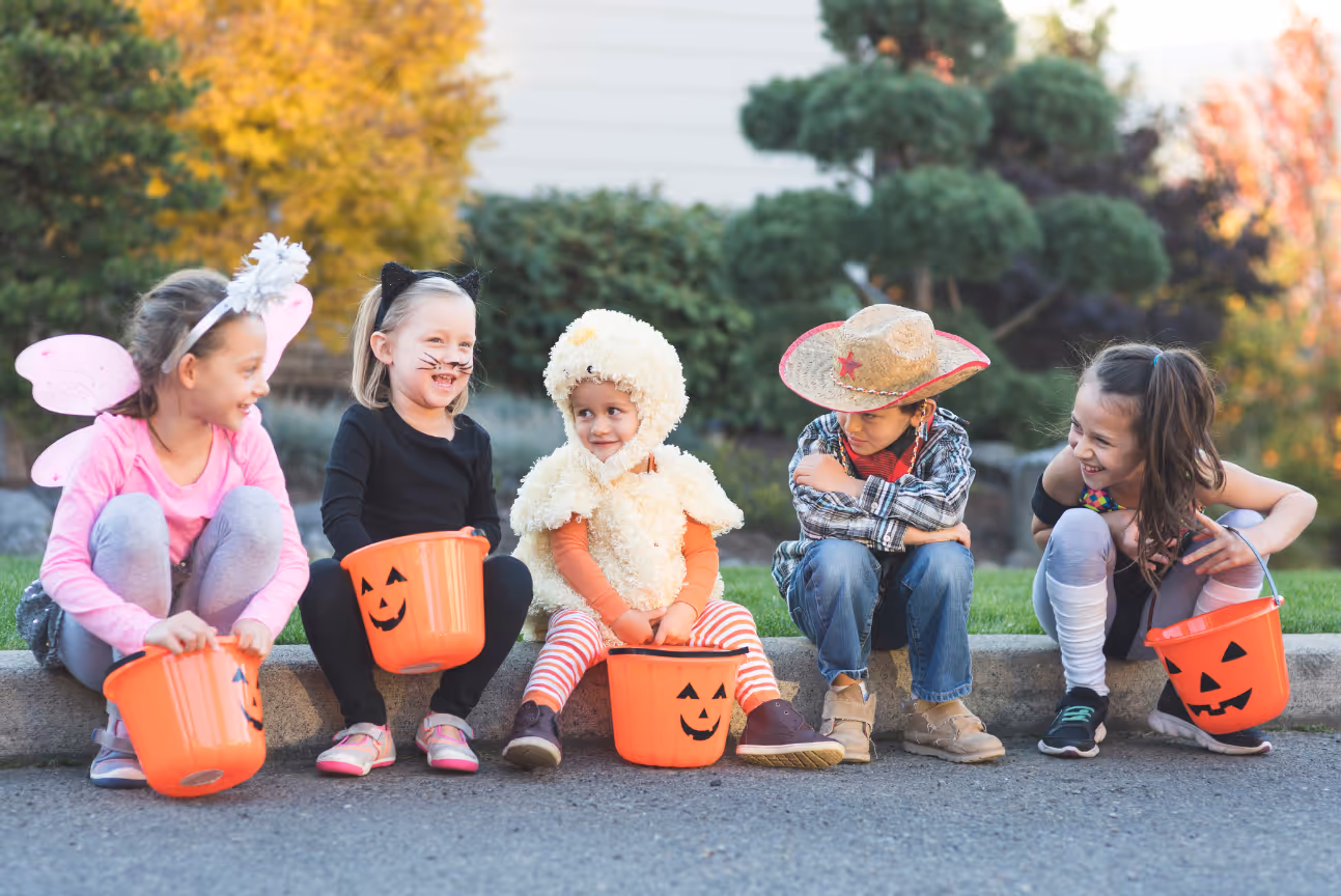 Five young children in Halloween costumes, such as a cat, fairy, duck, and cowboy, sit on a curb laughing and holding orange pumpkin buckets.