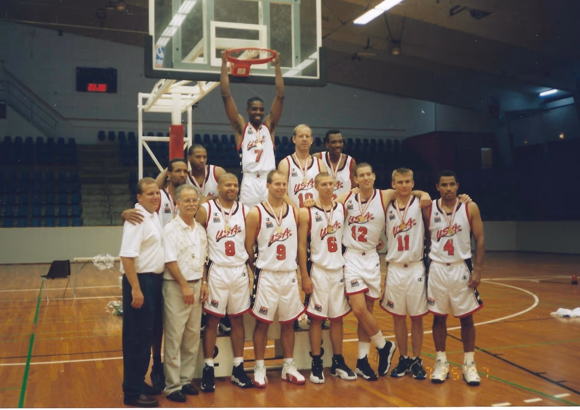 Team USA Deaf Men’s Basketball players and coaches pose under a hoop on a wooden court. One player hangs from the rim while others stand arm-in-arm wearing white USA uniforms and medals around their necks.