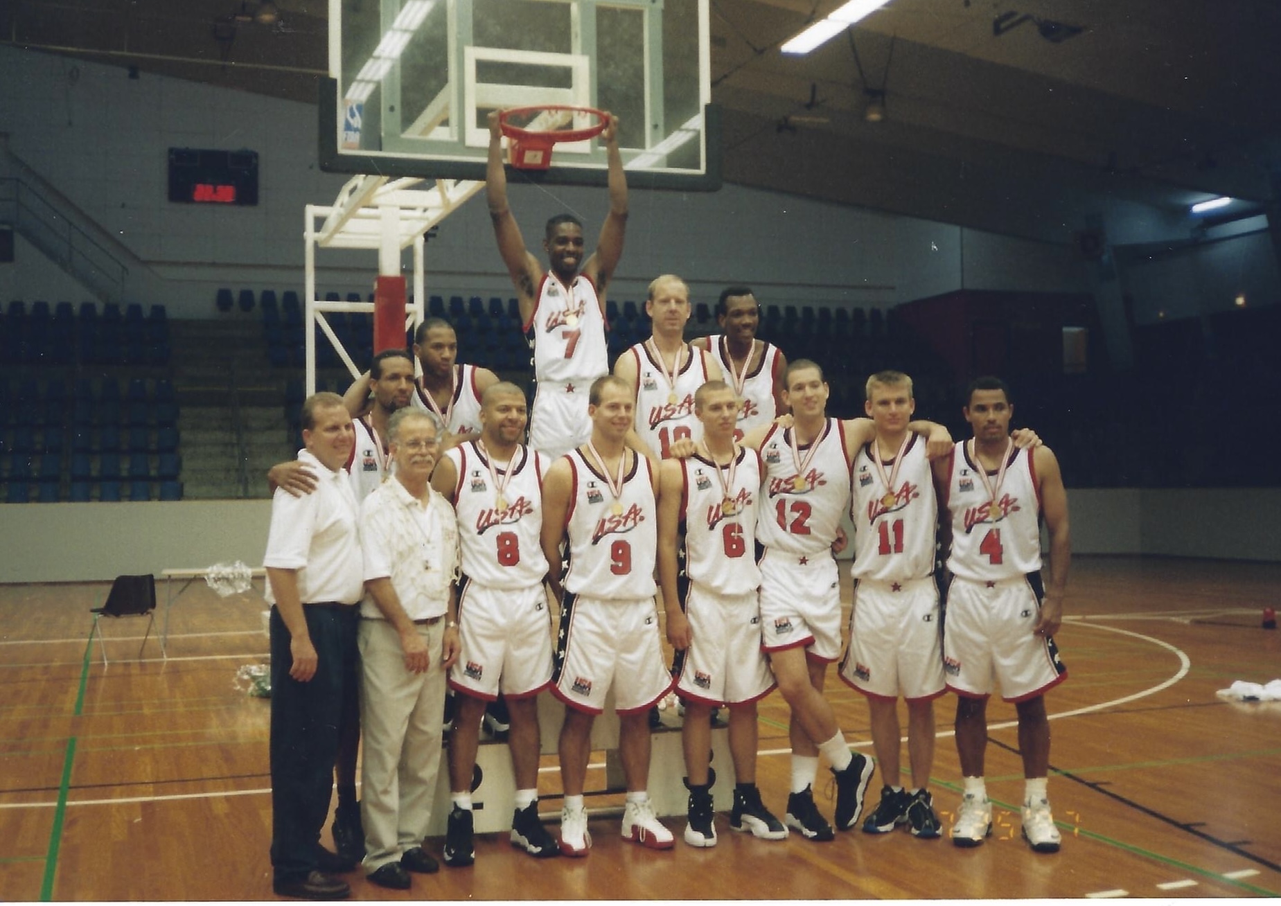 Team USA Deaf Men’s Basketball players and coaches pose under a hoop on a wooden court. One player hangs from the rim while others stand arm-in-arm wearing white USA uniforms and medals around their necks.