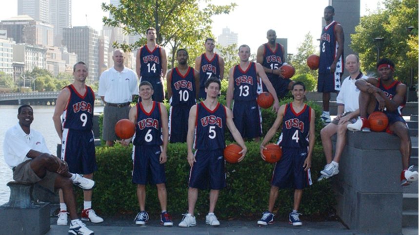 Team USA Deaf Men’s Basketball players and coaches pose outdoors near a waterfront city skyline. They wear navy blue USA uniforms and hold basketballs, standing and sitting together in a relaxed team photo.
