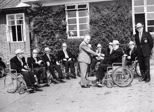 A black-and-white photo of Dr. Ludwig Guttmann shaking hands with a veteran in a wheelchair outside the Stoke Mandeville Hospital. Several men in wheelchairs line up beside them, smiling, marking one of the early moments in the development of the Paralympic movement.