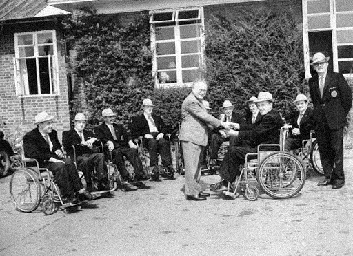 A black-and-white photo of Dr. Ludwig Guttmann shaking hands with a veteran in a wheelchair outside the Stoke Mandeville Hospital. Several men in wheelchairs line up beside them, smiling, marking one of the early moments in the development of the Paralympic movement.