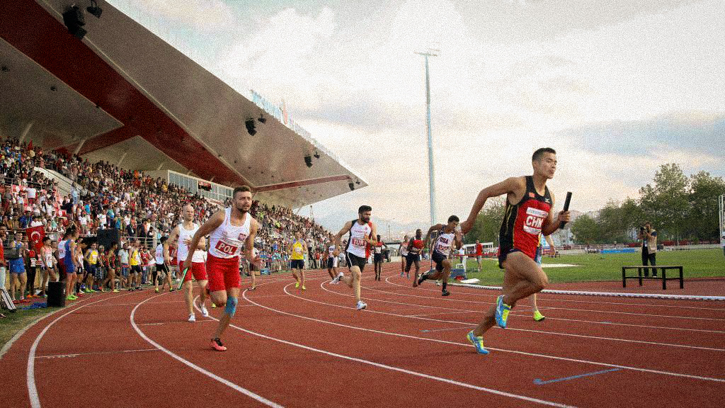 A group of Deaf track athletes from several countries sprint around a crowded stadium curve during a relay race at the Deaflympics. One runner from China accelerates ahead with the baton while athletes from Poland and other nations push forward behind him. The packed stands cheer under a sweeping red-and-white stadium roof.