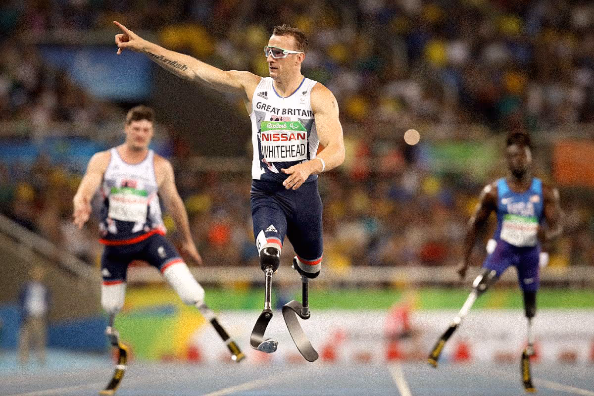 A Paralympic sprinter from Great Britain races ahead on carbon-fiber running blades, pointing toward the finish line with determination. Athletes from other countries follow behind him on the track, their own blade prostheses visible as they sprint in a packed stadium.
