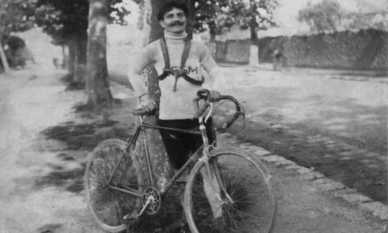 A historical black-and-white portrait of Eugène Rubens-Alcais standing outdoors beside his bicycle. Wearing a cycling jersey and sporting a proud smile, he appears along a tree-lined path, representing the early pioneering spirit that led to the founding of the Deaflympics.
