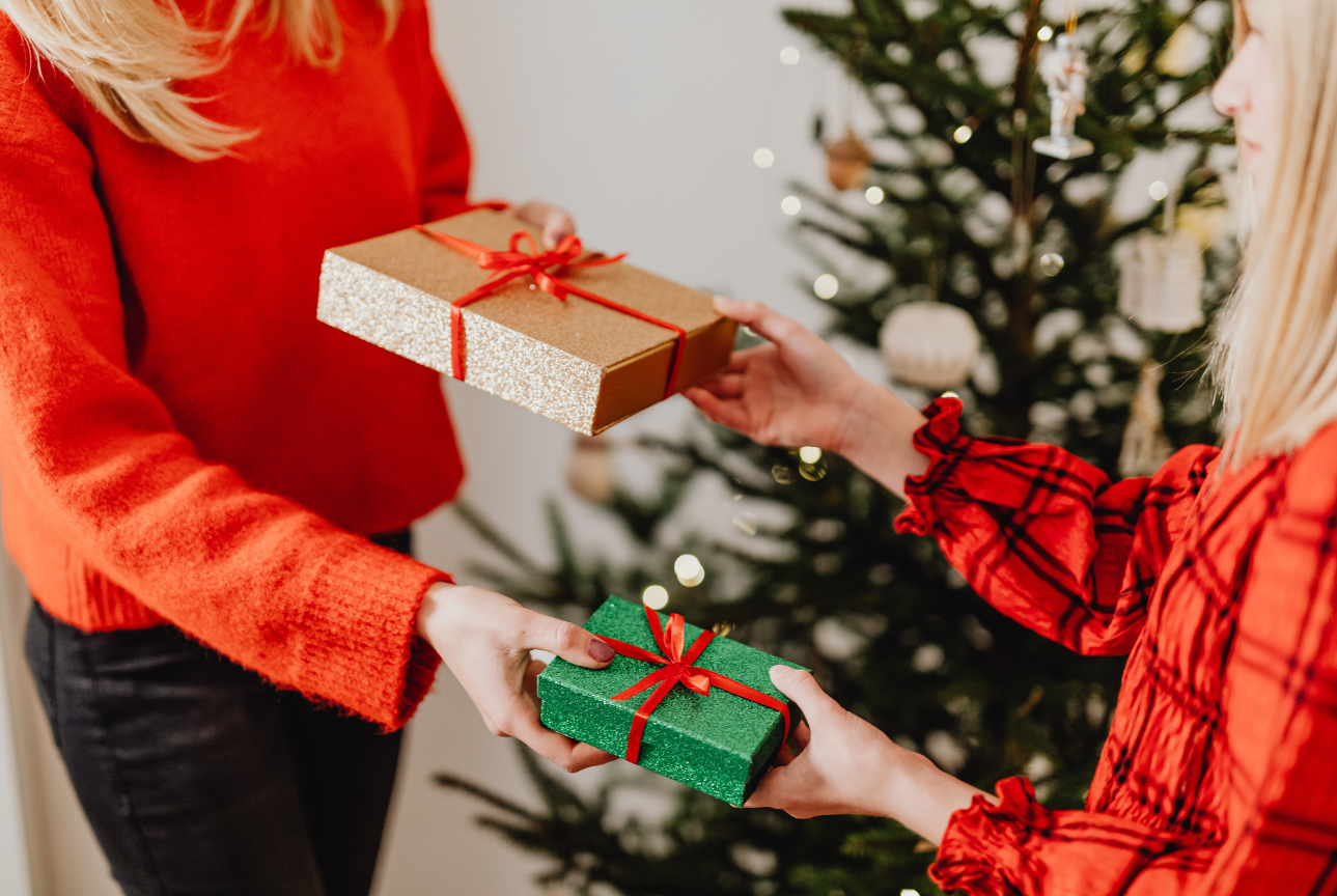 Two people in festive red sweaters exchange wrapped gifts in front of a decorated Christmas tree. One person hands over a gold box tied with a red ribbon while receiving a smaller green gift in return. Soft holiday lights glow in the background, creating a warm, celebratory atmosphere.