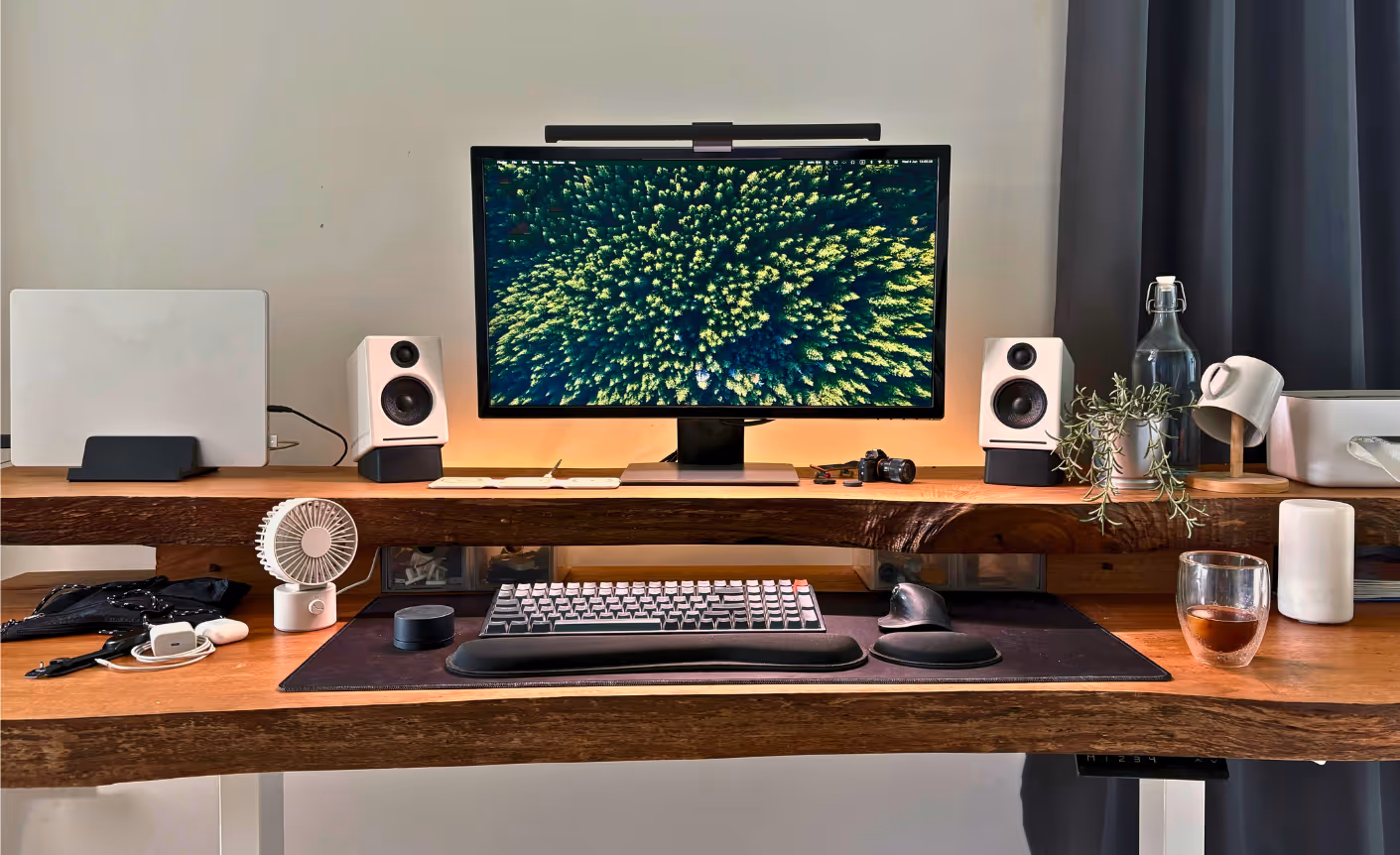 A neatly arranged home workspace with a large monitor displaying an aerial view of a dense green forest. The desk features white speakers, a keyboard with a wrist rest, a trackball mouse, a laptop on a stand, small plants, a camera, and a glass of tea. Warm lighting and natural wood textures create a calming, organized environment.