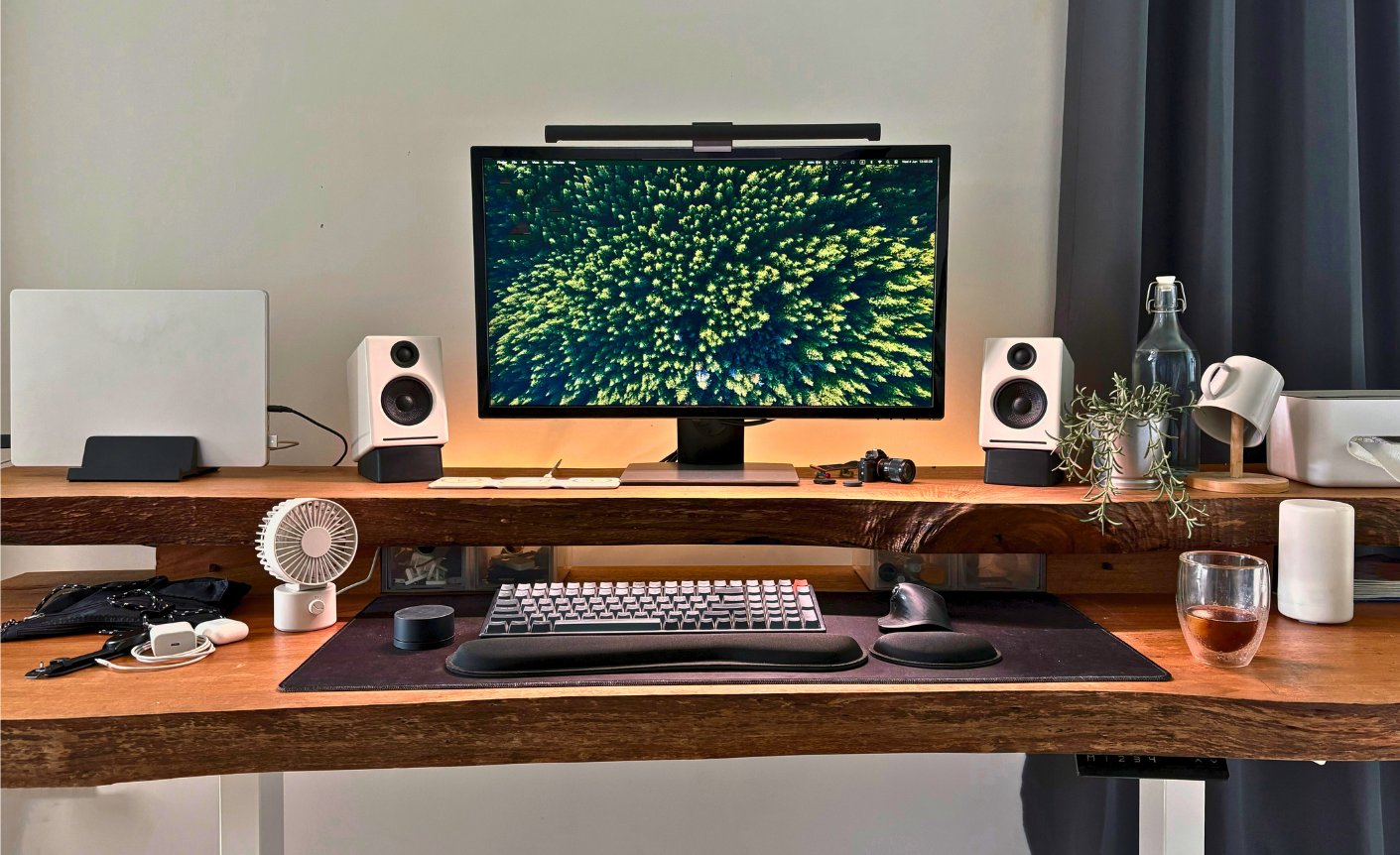 A neatly arranged home workspace with a large monitor displaying an aerial view of a dense green forest. The desk features white speakers, a keyboard with a wrist rest, a trackball mouse, a laptop on a stand, small plants, a camera, and a glass of tea. Warm lighting and natural wood textures create a calming, organized environment.