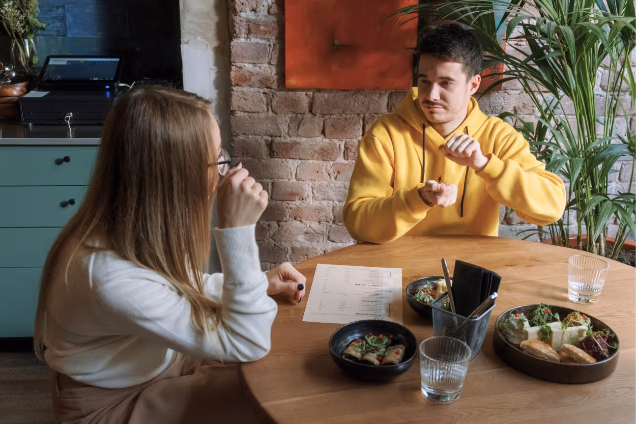 Two people sit at a restaurant table, signing to each other while sharing a meal. The man signs with both hands as the woman watches attentively, reflecting a natural ASL conversation in a casual setting.