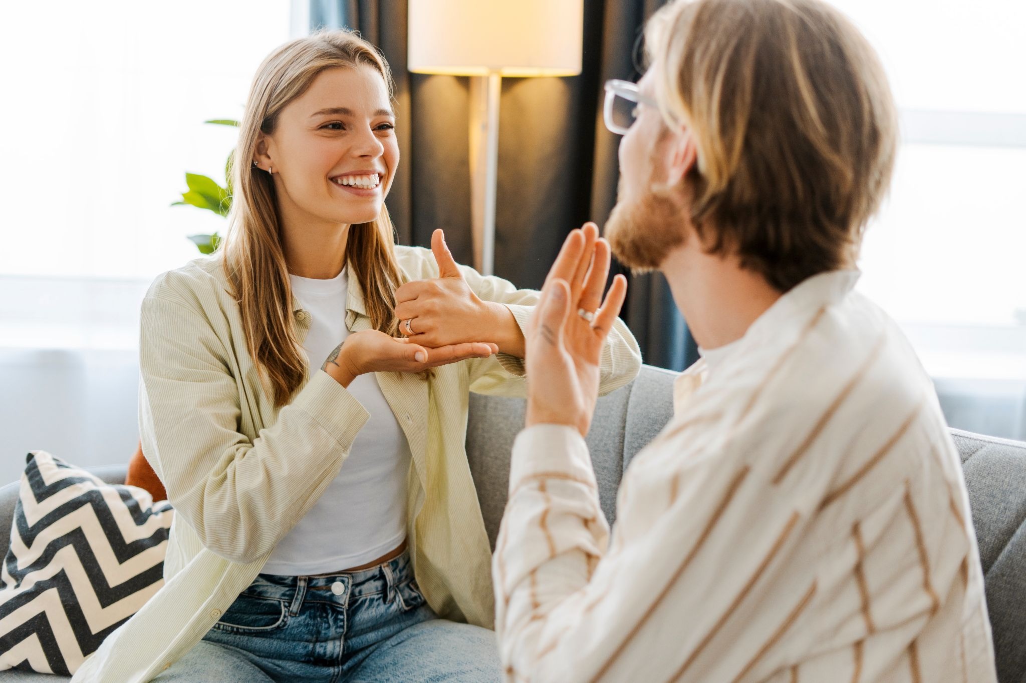 A woman and a man sit on a couch facing each other, smiling as they have a friendly conversation in American Sign Language. Both are engaged and expressive, creating a warm, relaxed learning environment.