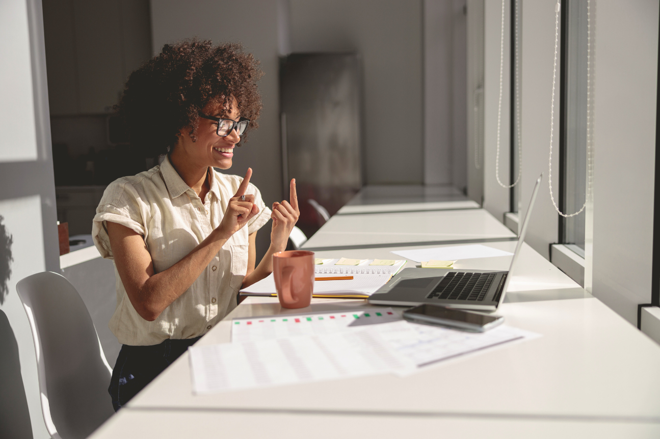 A woman sits at a bright desk during a video call, smiling as she signs toward her laptop. Papers, a notebook, and a mug sit nearby, creating a focused and welcoming workspace.