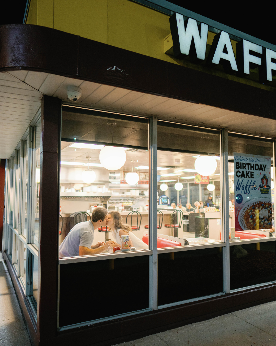 A couple shares a kiss while sitting inside a Waffle House diner at night, visible through the restaurant’s front window during an engagement session.