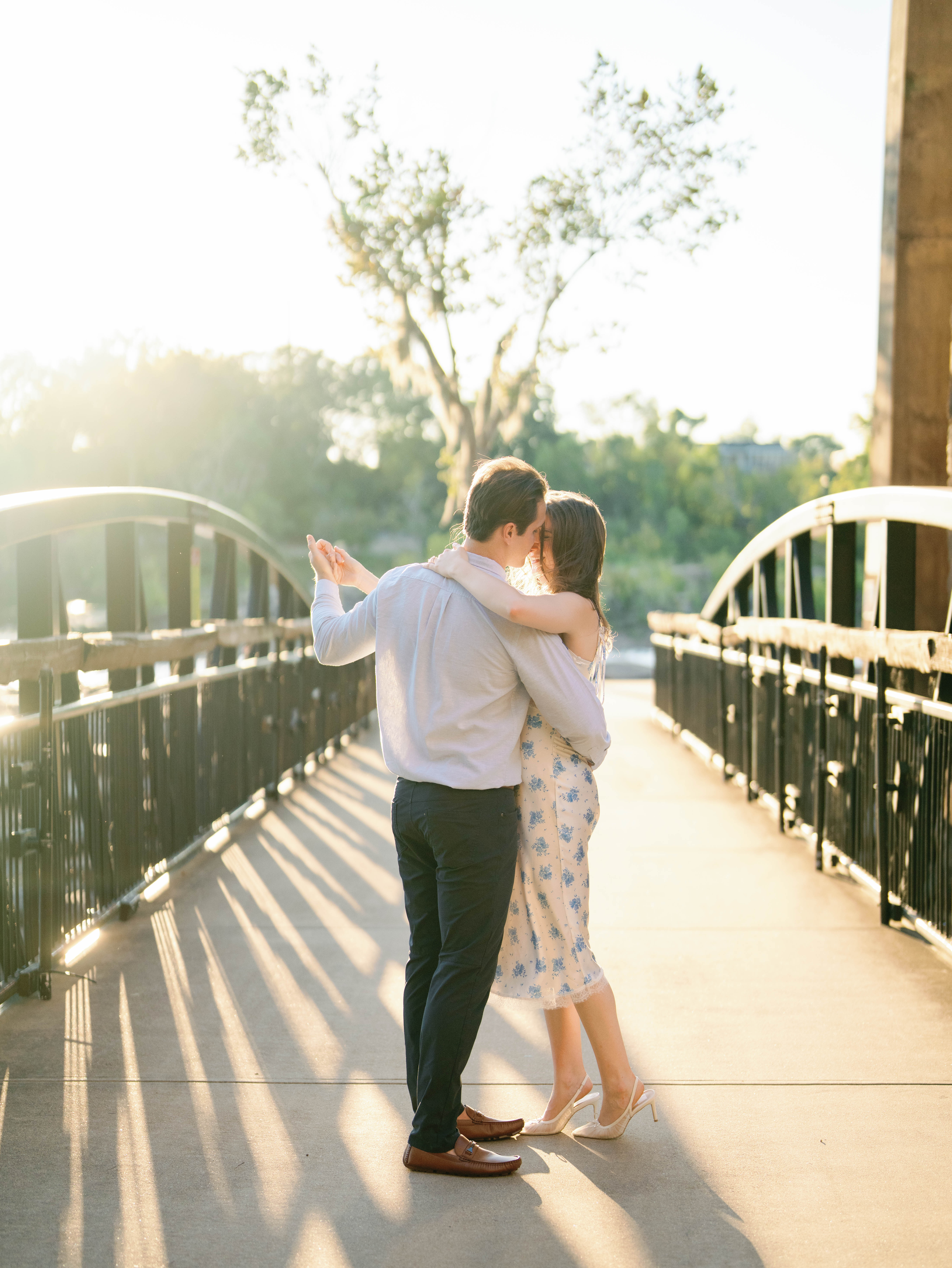 A couple embraces and dances on a pedestrian bridge at golden hour, surrounded by soft sunlight and greenery during an engagement photo shoot.