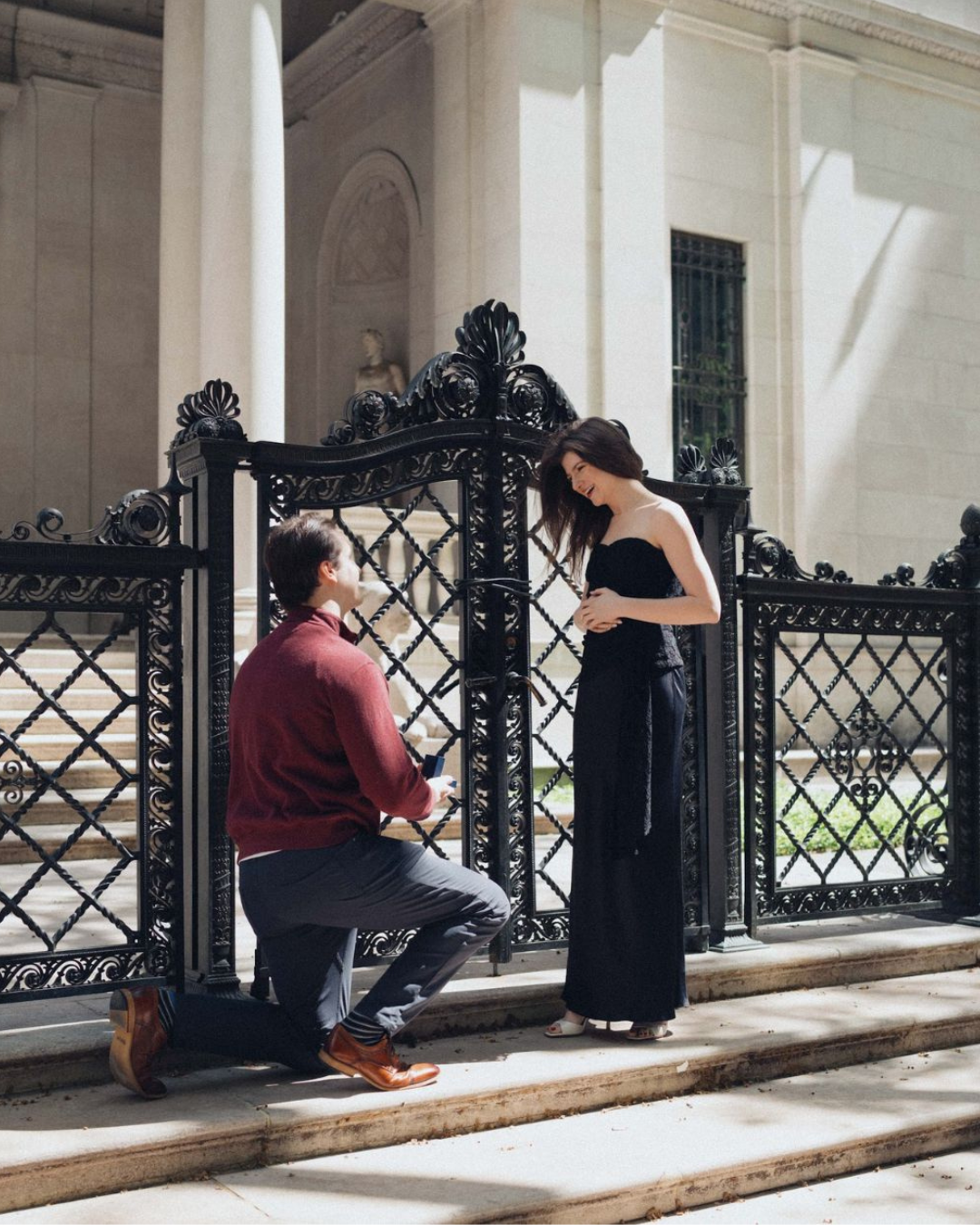 A man kneels and proposes to his partner in front of an ornate iron gate and classical building in New York City during a surprise engagement moment.