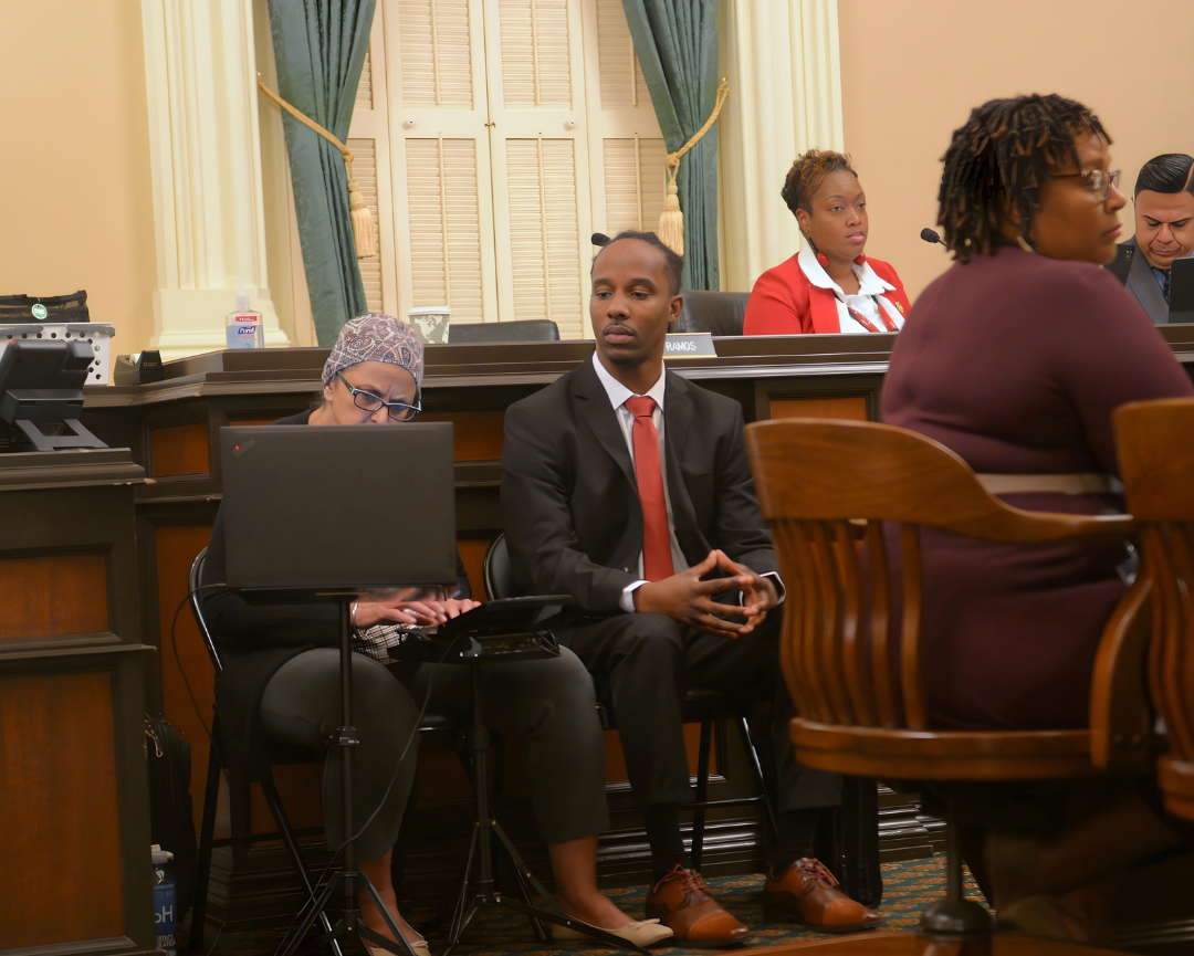 Tremmel Watson sits attentively in a formal, wood-paneled hearing room next to a CART provider. The captioner is focused on typing on a steno machine, while Tremmel looks forward, illustrating the use of live human captioning for communication access during a professional meeting.