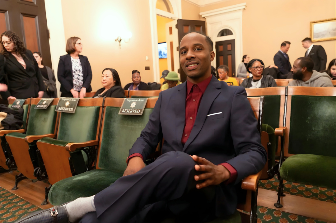Tremmel Watson, a Black man dressed in a navy blue suit and burgundy dress shirt, sits confidently with his legs crossed in a formal legislative chamber. He is seated in a row of green velvet chairs marked with "RESERVED" signs, looking directly at the camera.