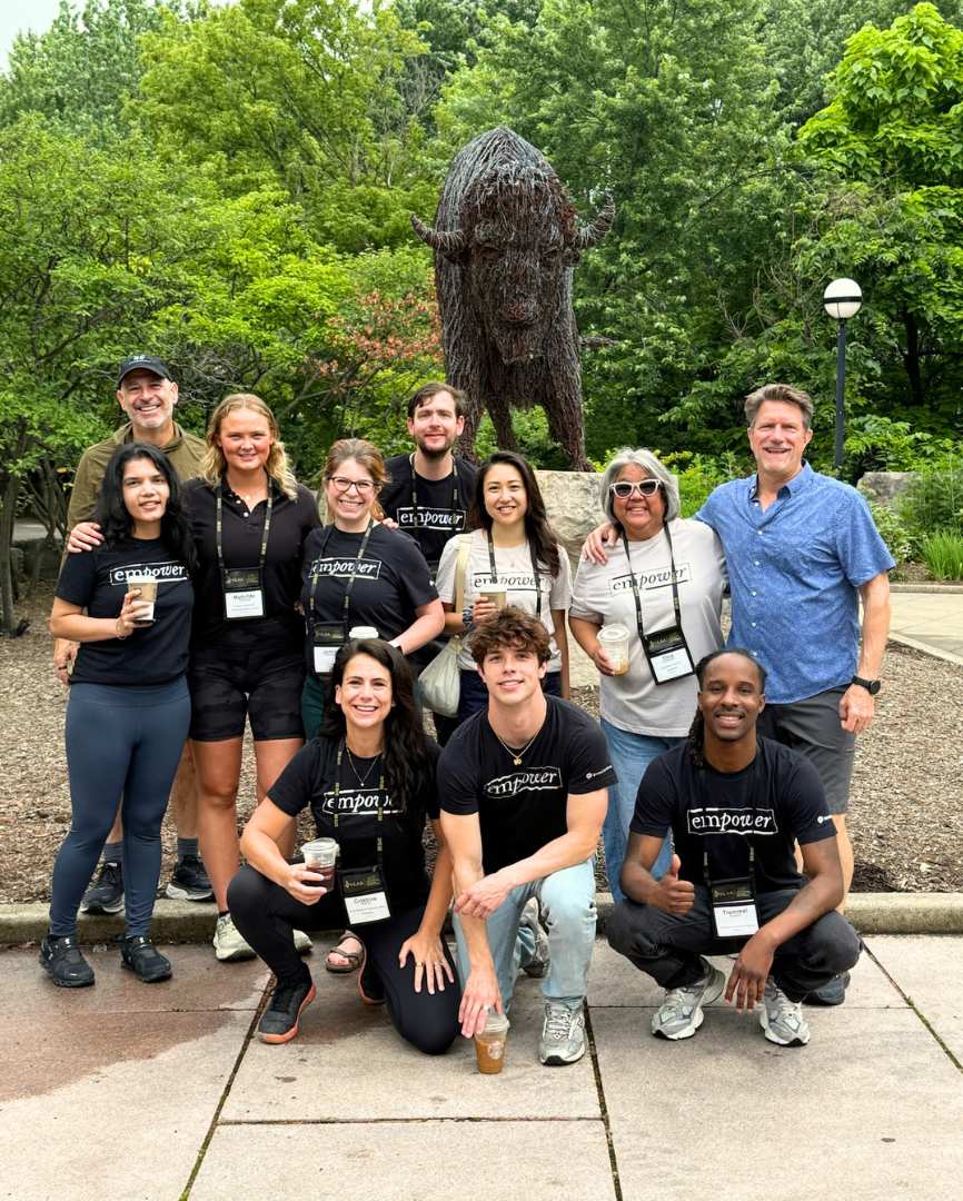 A group photograph of eleven smiling people posing outdoors on a concrete path in a wooded area. Most are wearing black T-shirts with "EMPOWER" printed on the front, a few wear light gray versions, and several wear name tag lanyards. The group is arranged with three people crouching in the front row and eight standing behind them. Many are holding disposable coffee-style cups, and the man crouching on the front right gives a thumbs-up gesture. Behind the group is a large, central statue of a bison made of textured, woven branches on a stone plinth, set against a background of green trees and foliage.
