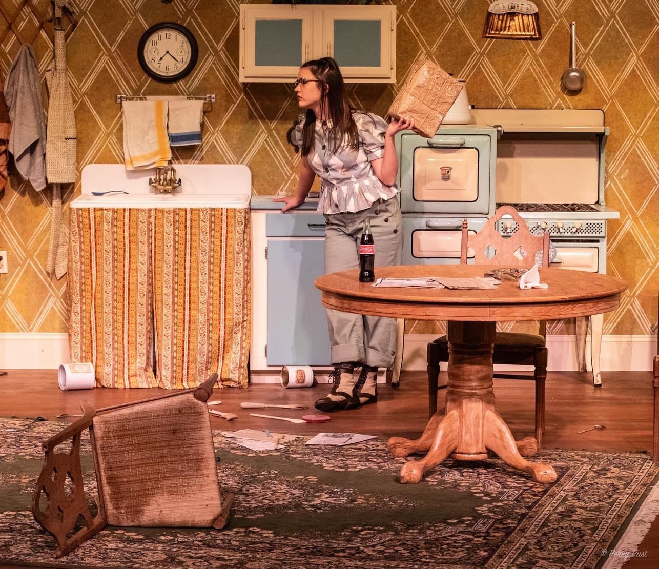 A dramatic stage scene set in a vintage-style kitchen with diamond-patterned wallpaper. Abigail, wearing a patterned blouse and trousers, stands tensely by a table, looking off-stage with a wary, focused expression while holding a brown wrapped package. In the foreground, a wooden chair lies overturned on a rug, suggesting a struggle or a moment of high tension.