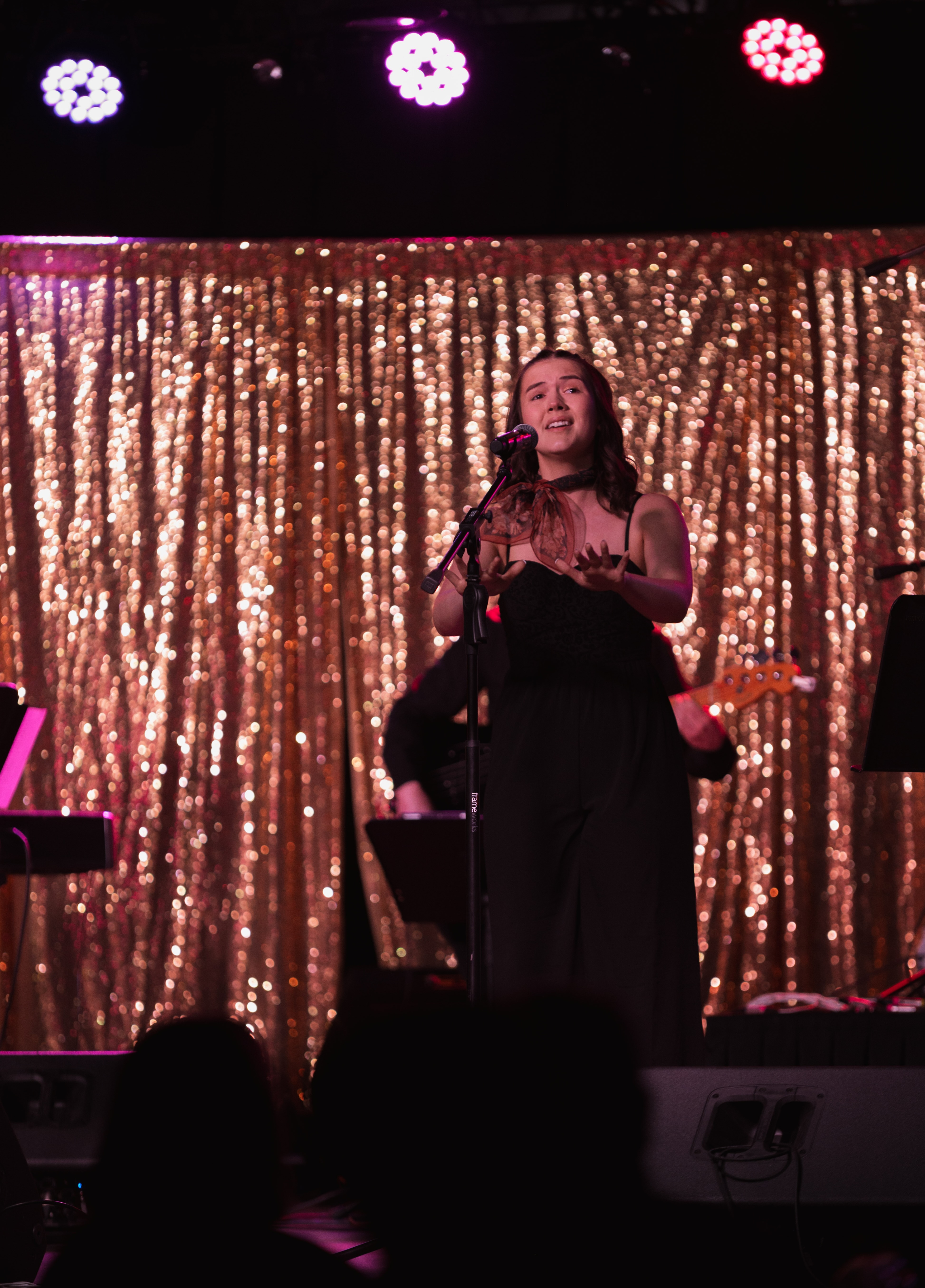 Abigail stands at a microphone on a stage lit with moody purple and red lights. She is wearing a black jumpsuit and is captured mid-sign, using ASL to perform. The background is a shimmering, gold sequined curtain that catches the stage lights.