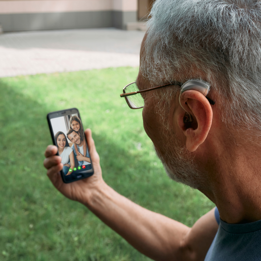 An older man wearing a behind-the-ear hearing aid uses a smartphone for a video call with a smiling family outdoors.