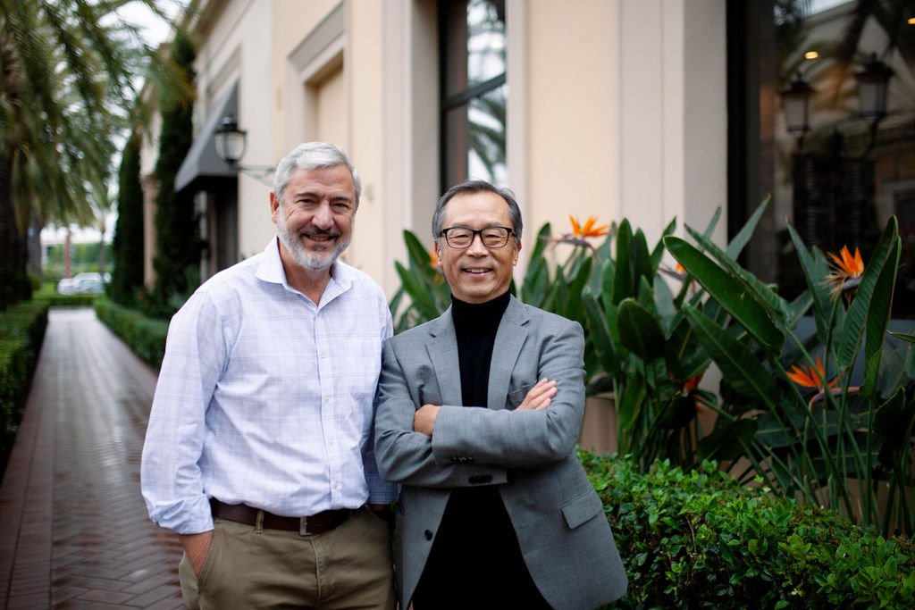Two professionally dressed older men stand side-by-side on an outdoor paver walkway, both smiling. The man on the left wears a plaid shirt and the man on the right wears a blazer and turtleneck with arms crossed. Behind them is an upscale building with large windows, surrounded by green hedges and bird of paradise flowers.