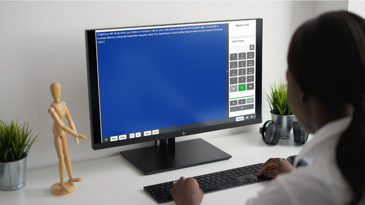An over-the-shoulder view of a woman typing on a keyboard at a white desk, looking at a computer monitor. The screen displays the InnoCaption DeskView interface, featuring a large blue call captioning window and a digital dialer keypad.