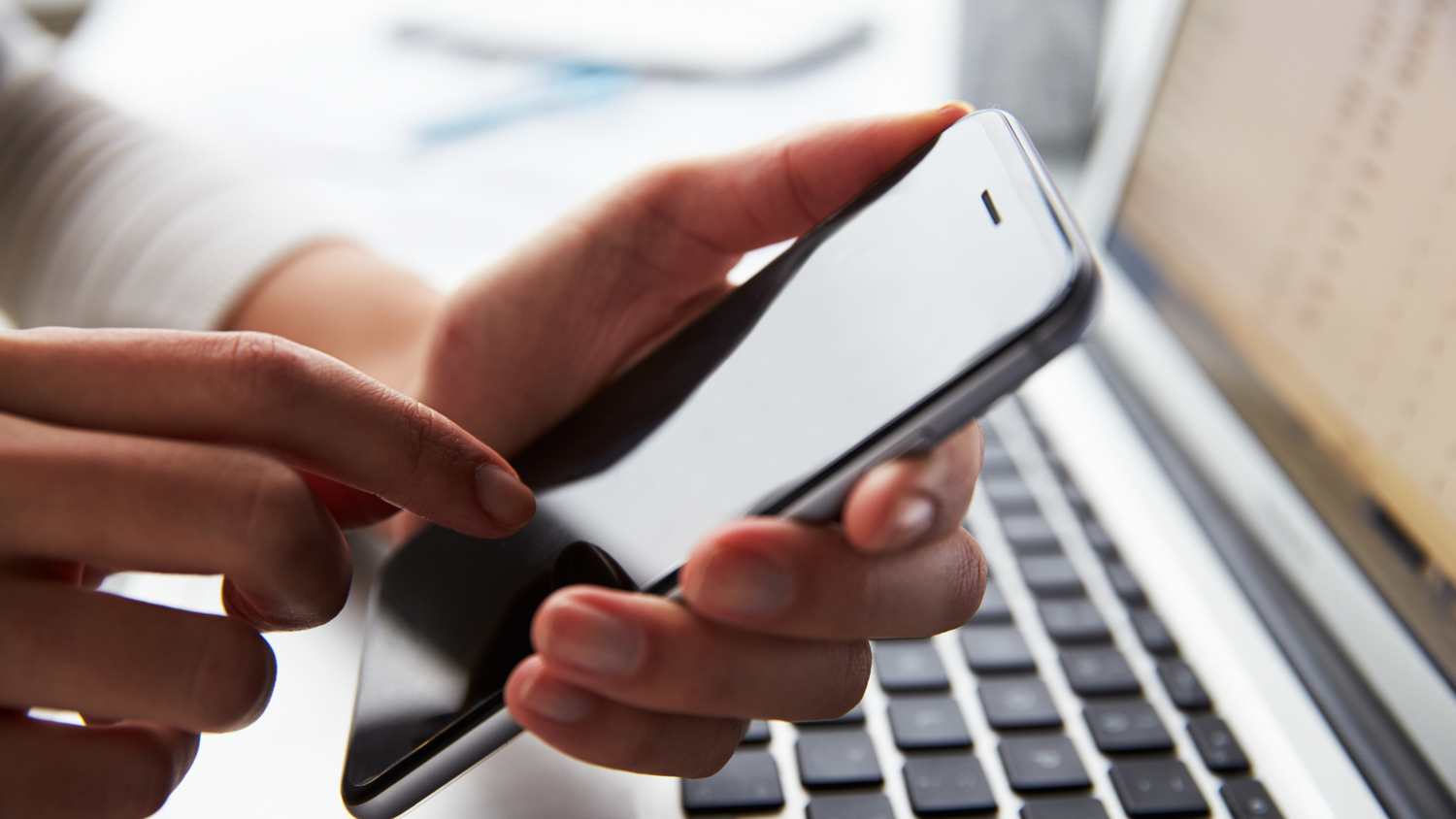 alt="A close-up of a person's hands holding a smartphone and tapping the screen, next to an open laptop on a white desk.