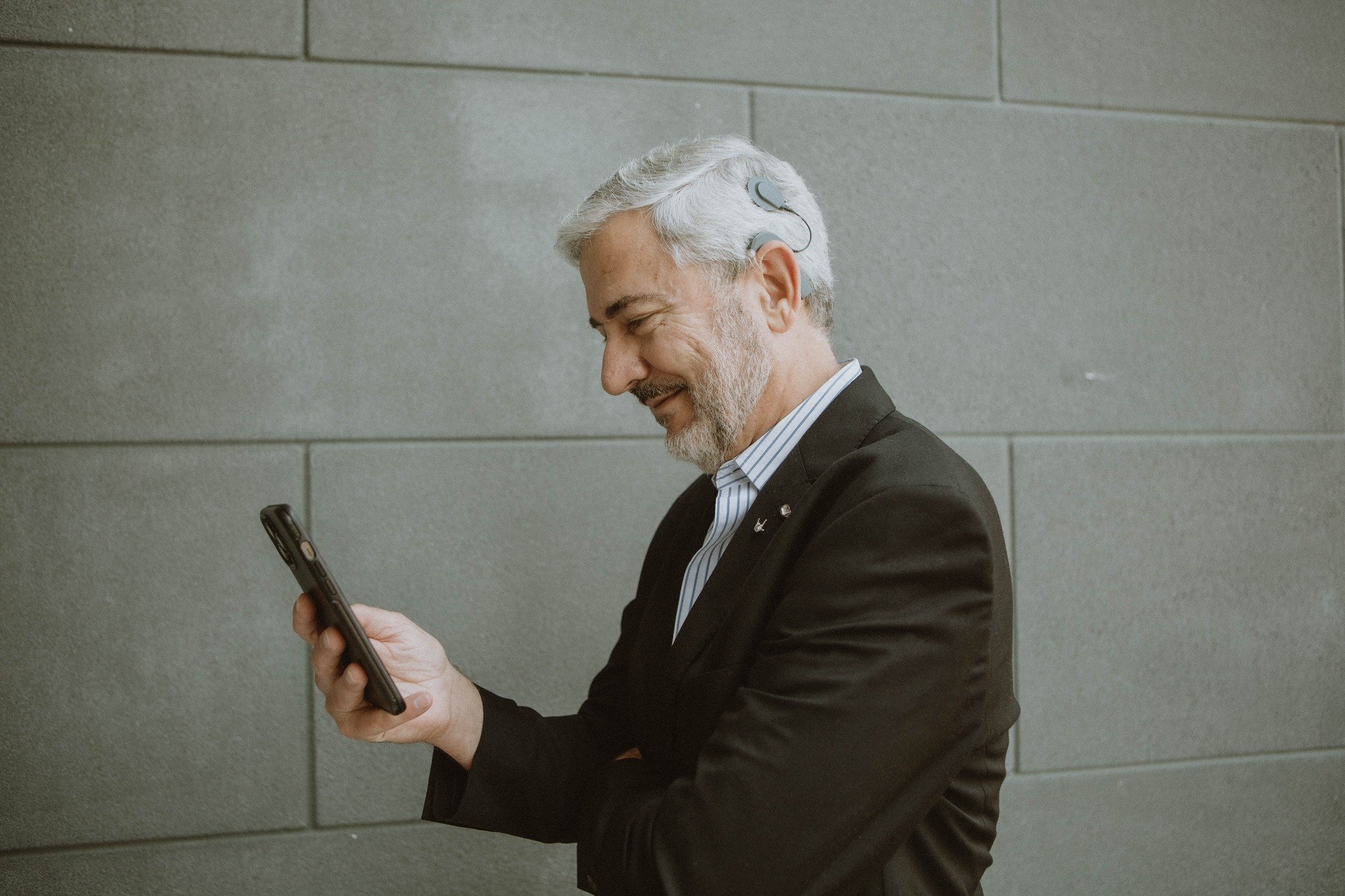 A side profile of InnoCaption Co-CEO Joseph Duarte looking down at a smartphone. He has grey hair and a beard, and wears a black suit jacket and a cochlear implant against a grey tiled wall background.
