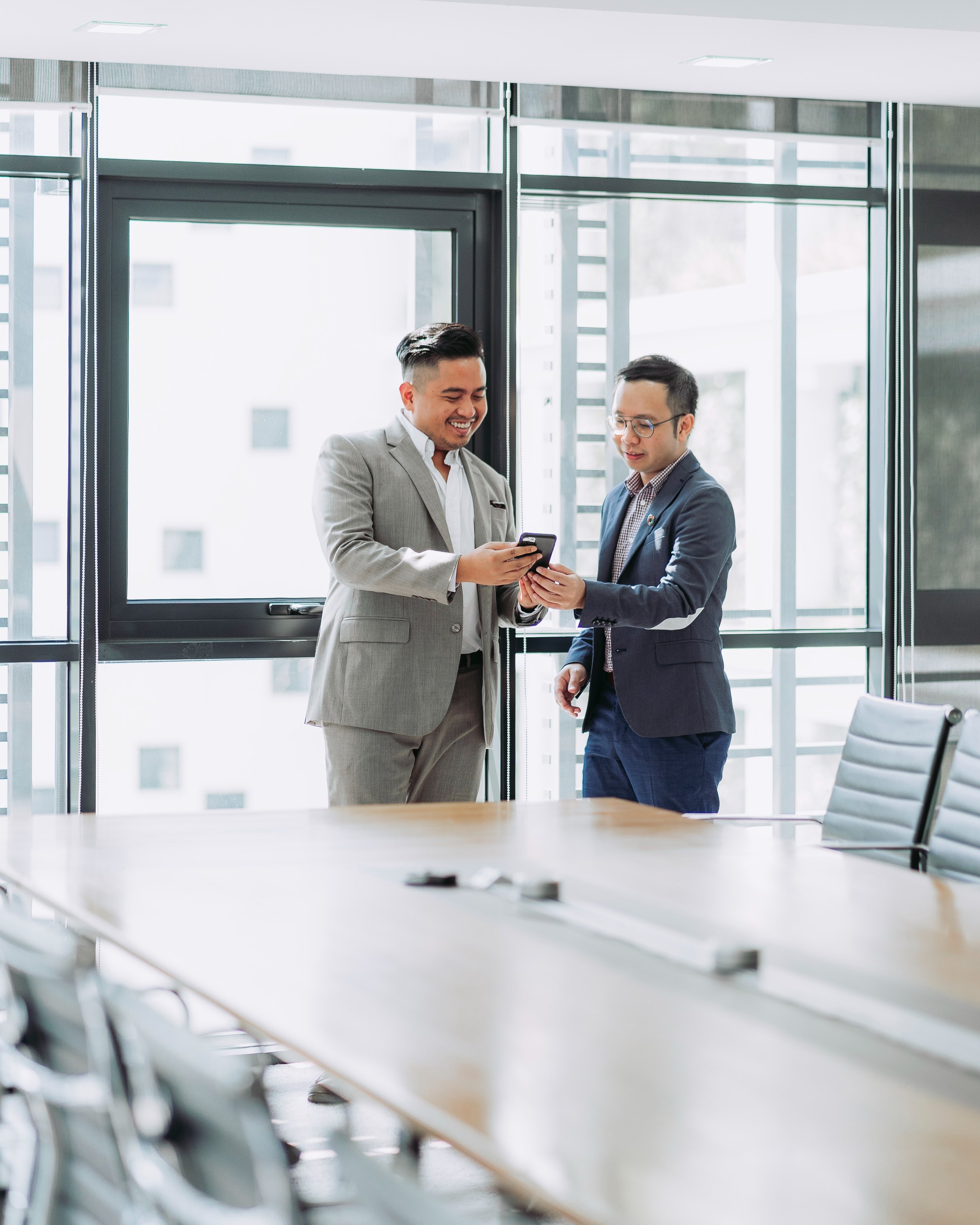 Two smiling businessmen in professional suits stand in a modern conference room looking at a smartphone together. A long wooden conference table is in the foreground with large floor-to-ceiling windows in the background.