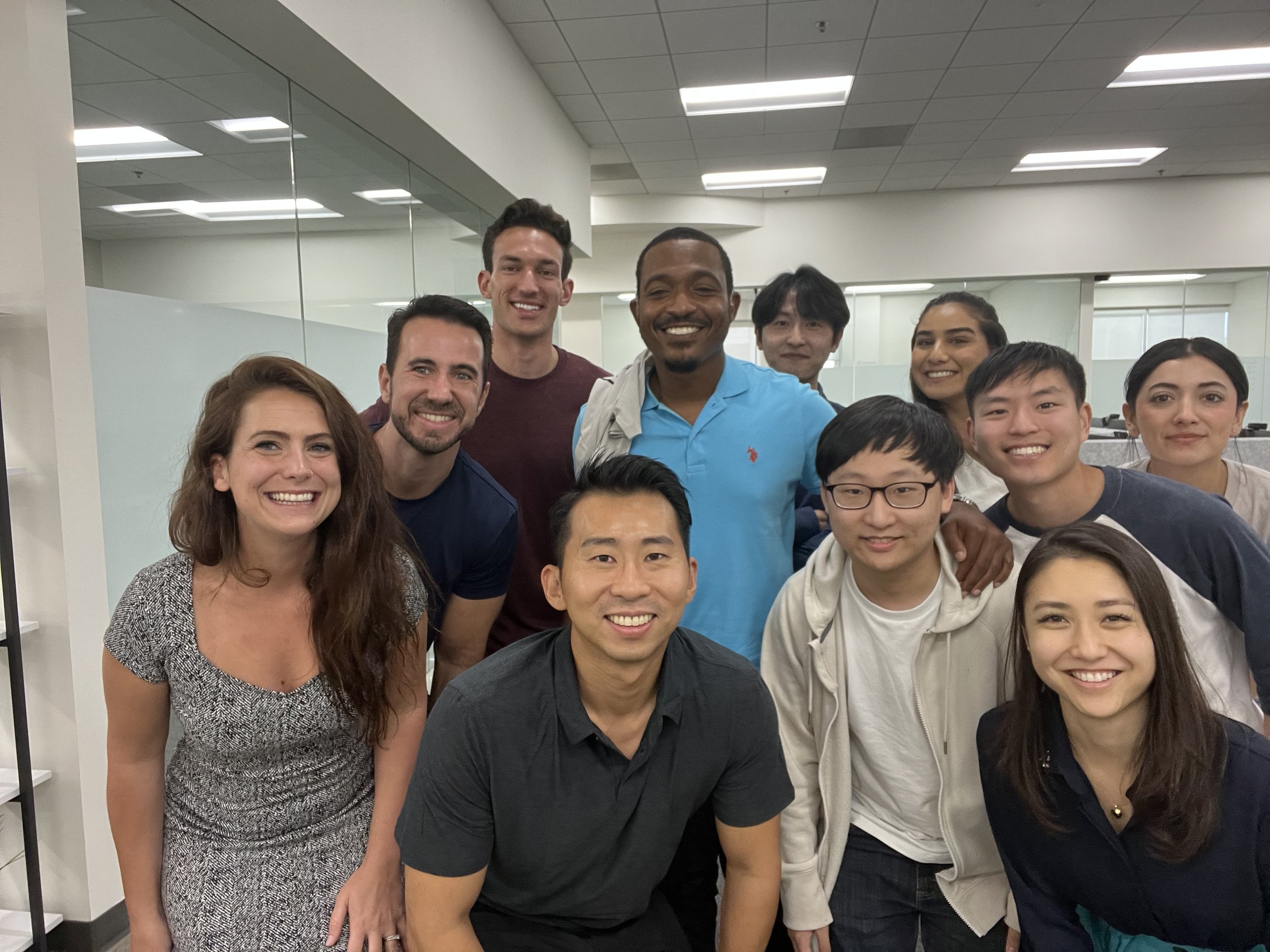 A diverse group of ten smiling people, including John Autry II in a bright blue polo shirt, posing together for a group photo inside a bright, modern office with glass walls.
