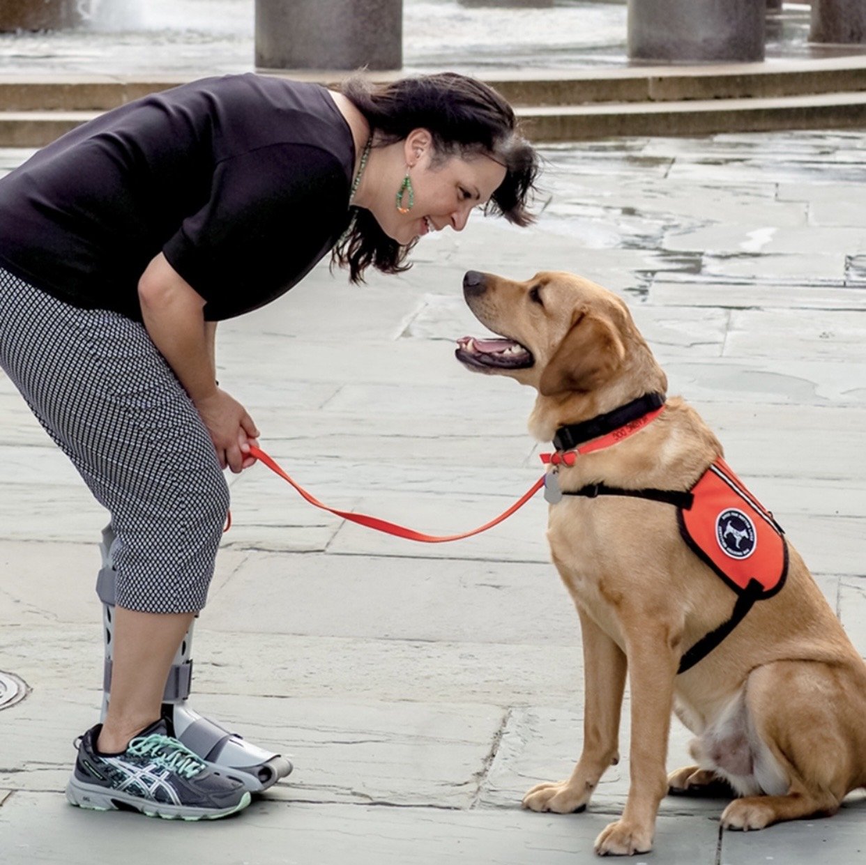 alt="A woman leans forward to smile at a yellow Labrador retriever sitting on a stone plaza. The dog is a hearing assistance service dog wearing a bright orange vest and looking up at her happily.