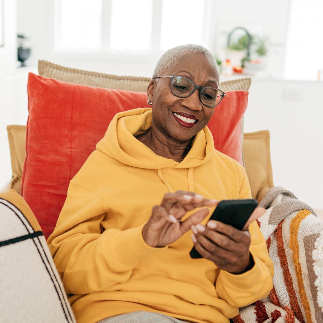 A smiling senior Black woman with short grey hair and glasses sits comfortably on a couch while using a smartphone. She is wearing a bright yellow hoodie and leaning against a large orange-red pillow.