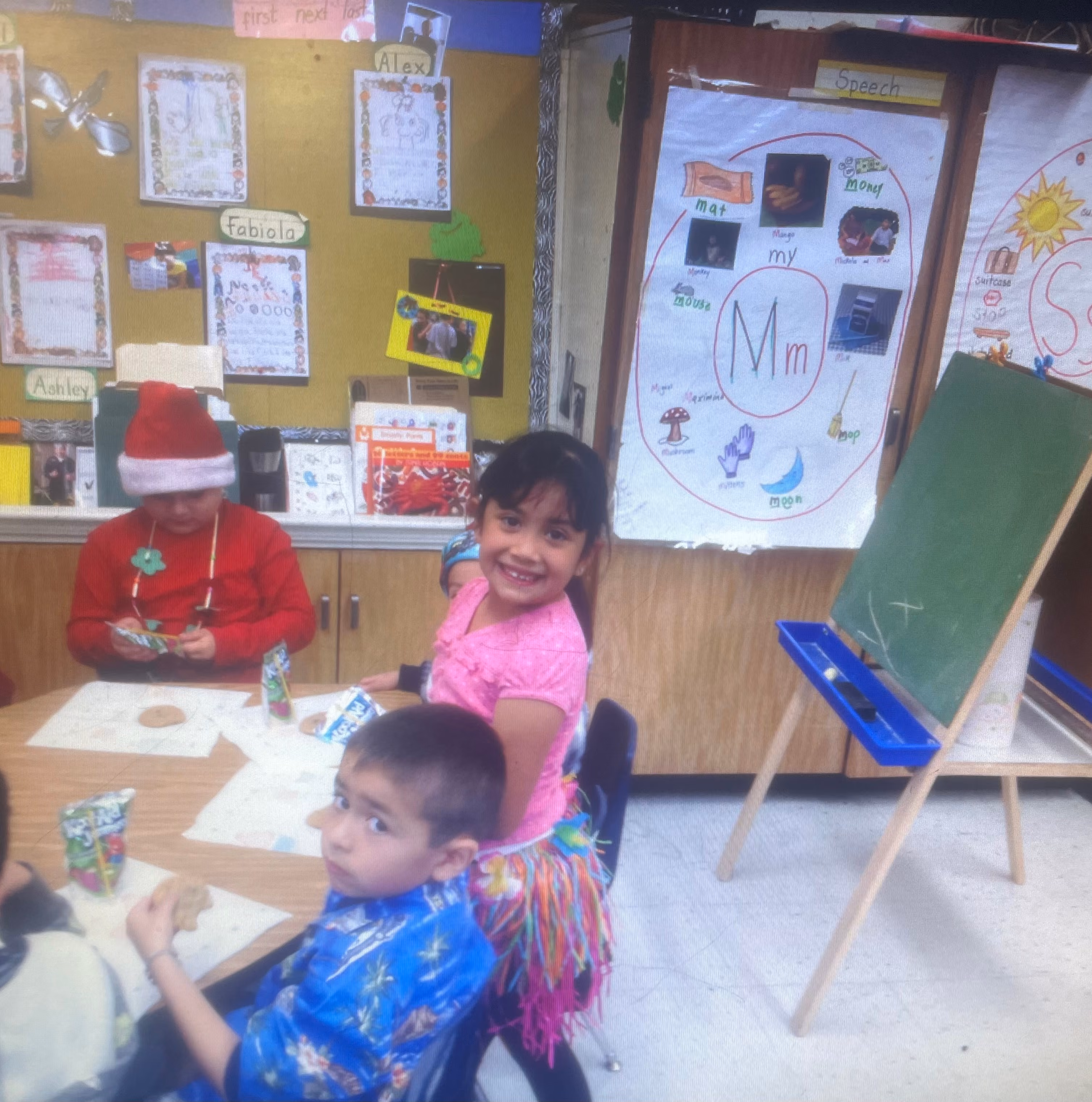 Fabiola Ramirez as a young child, about five years old, smiling over her shoulder at a classroom table during a speech session, with her name posted on the wall behind her beside a letter learning poster.
