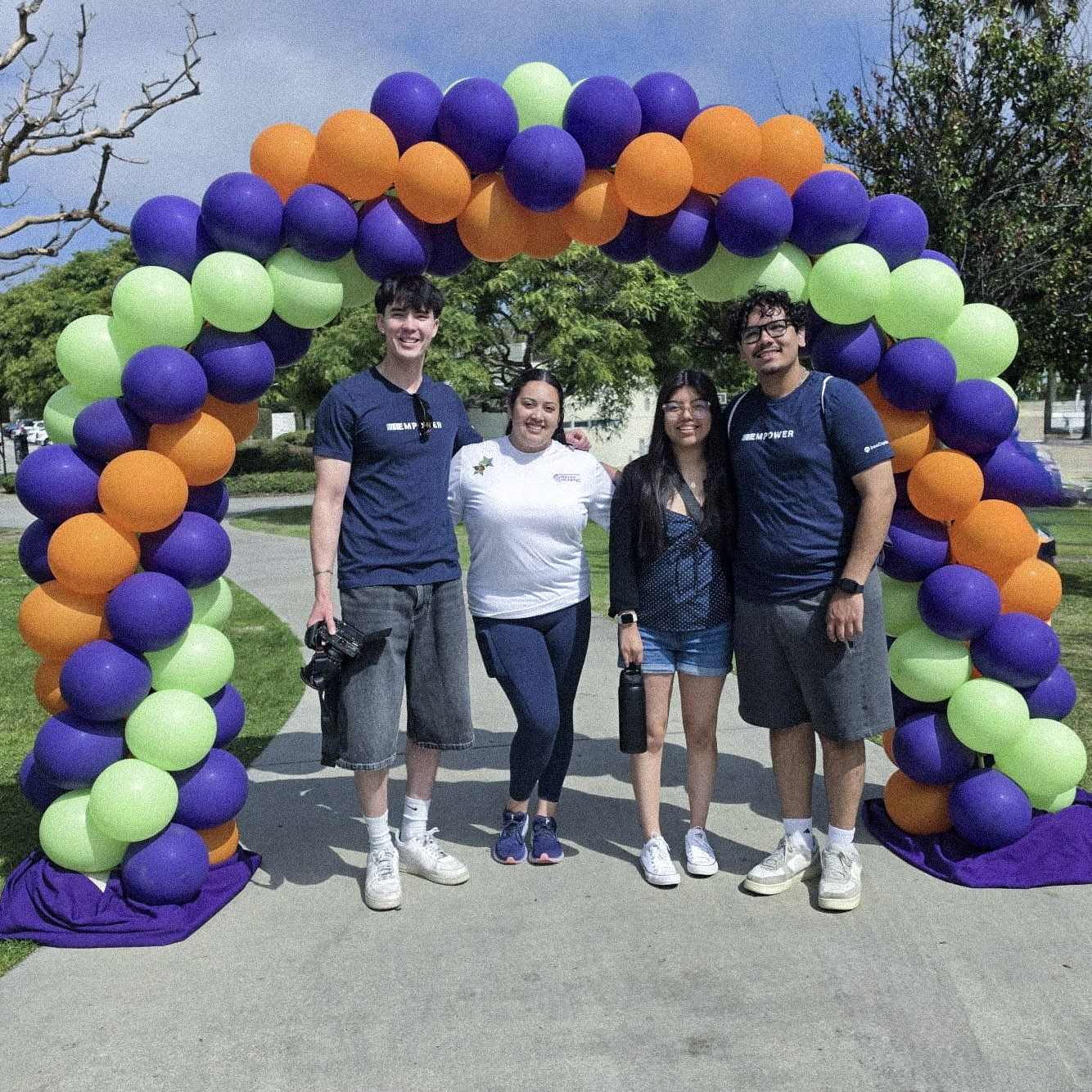 Fabiola Ramirez (second from right) stands with friends under a purple, green, and orange balloon arch at the HLAA Walk4Hearing, including Christian (far right), with two of the group wearing navy InnoCaption "Empower" shirts.