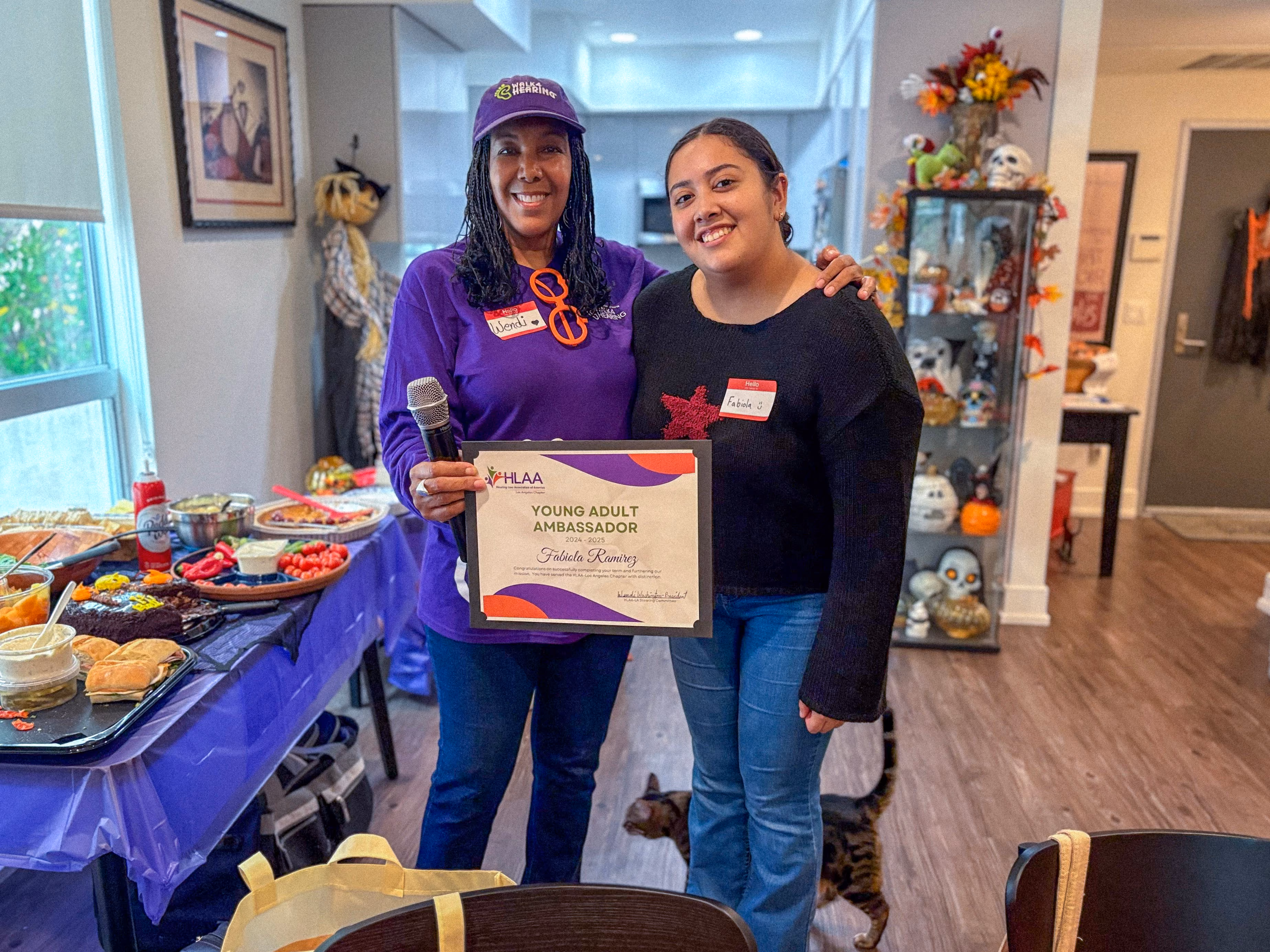 Fabiola Ramirez smiles next to HLAA Los Angeles chapter president Wendi Washington, who wears a Walk4Hearing cap, as the two hold Fabiola's 2024 to 2025 Young Adult Ambassador certificate.