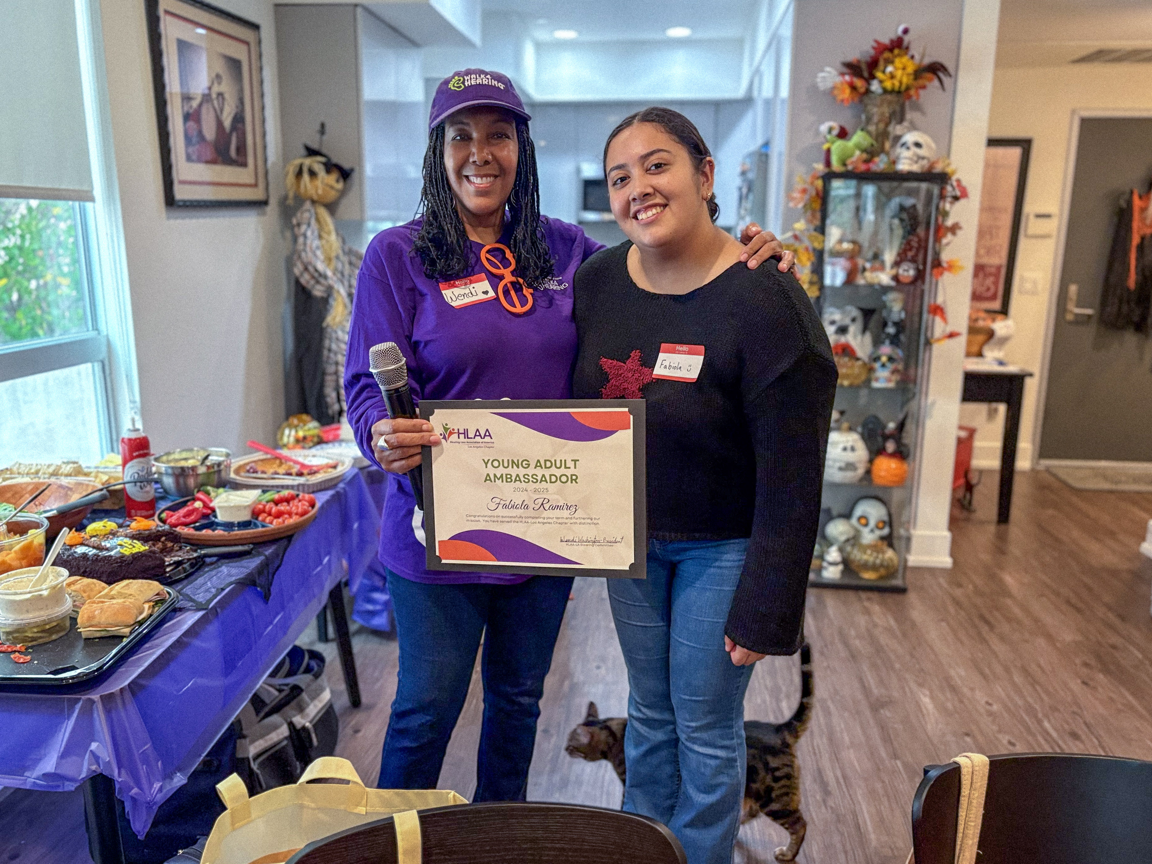 Fabiola Ramirez smiles next to HLAA Los Angeles chapter president Wendi Washington, who wears a Walk4Hearing cap, as the two hold Fabiola's 2024 to 2025 Young Adult Ambassador certificate.