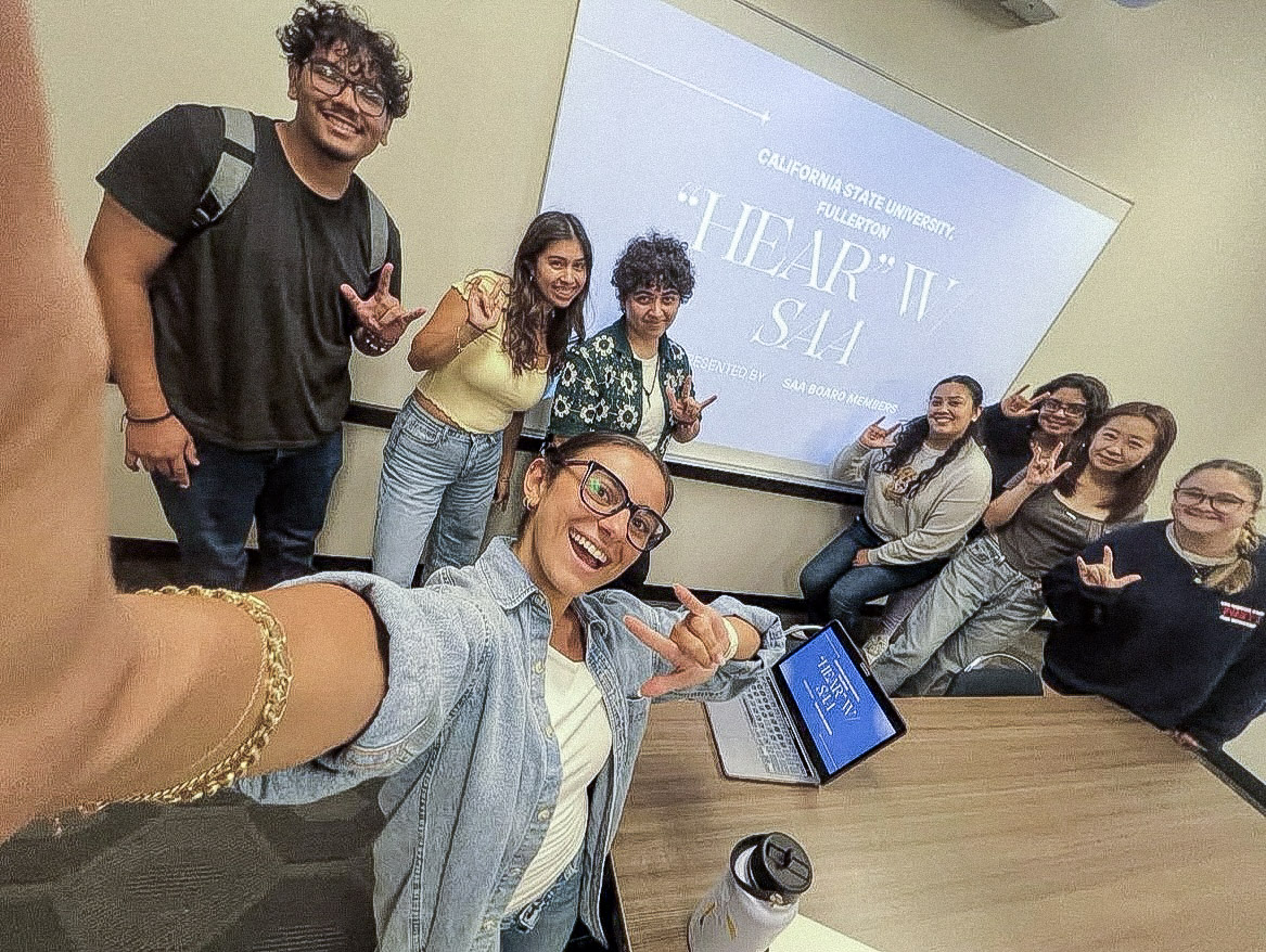 Fabiola Ramirez takes a group selfie with seven smiling friends, several flashing the ASL "I love you" sign, in front of a presentation screen reading "California State University, Fullerton, 'HEAR' w/ SAA, presented by SAA Board Members."