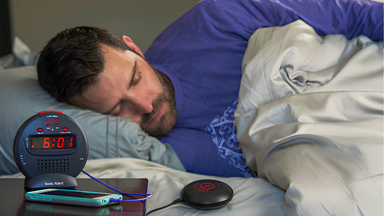 A Sonic Bomb alarm clock and bed shaker sit on a nightstand beside a sleeping person. The alarm clock display reads 6:01.