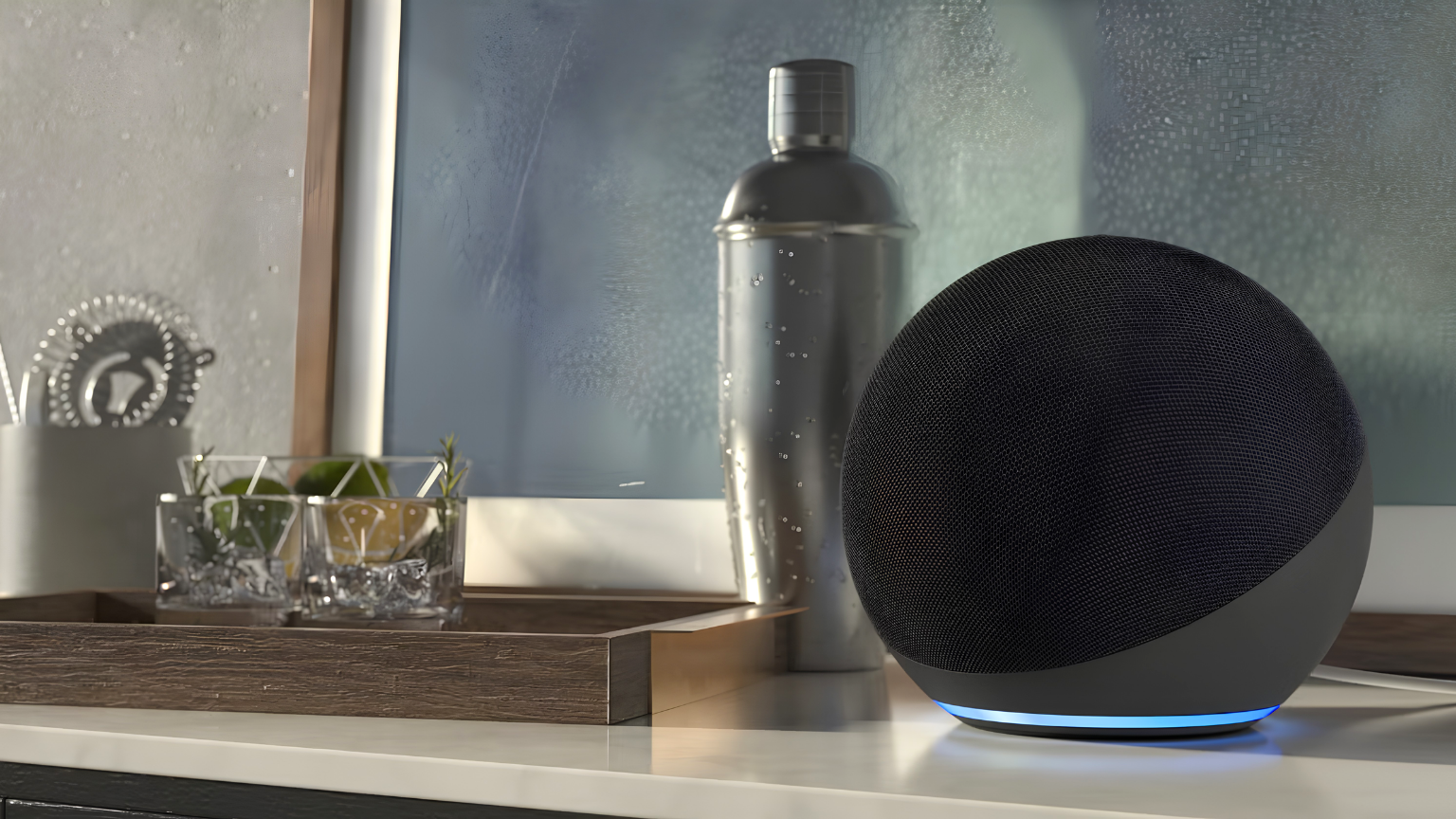 Amazon Echo smart speaker with a blue light ring sits on a kitchen counter beside glasses, citrus, and a cocktail shaker.