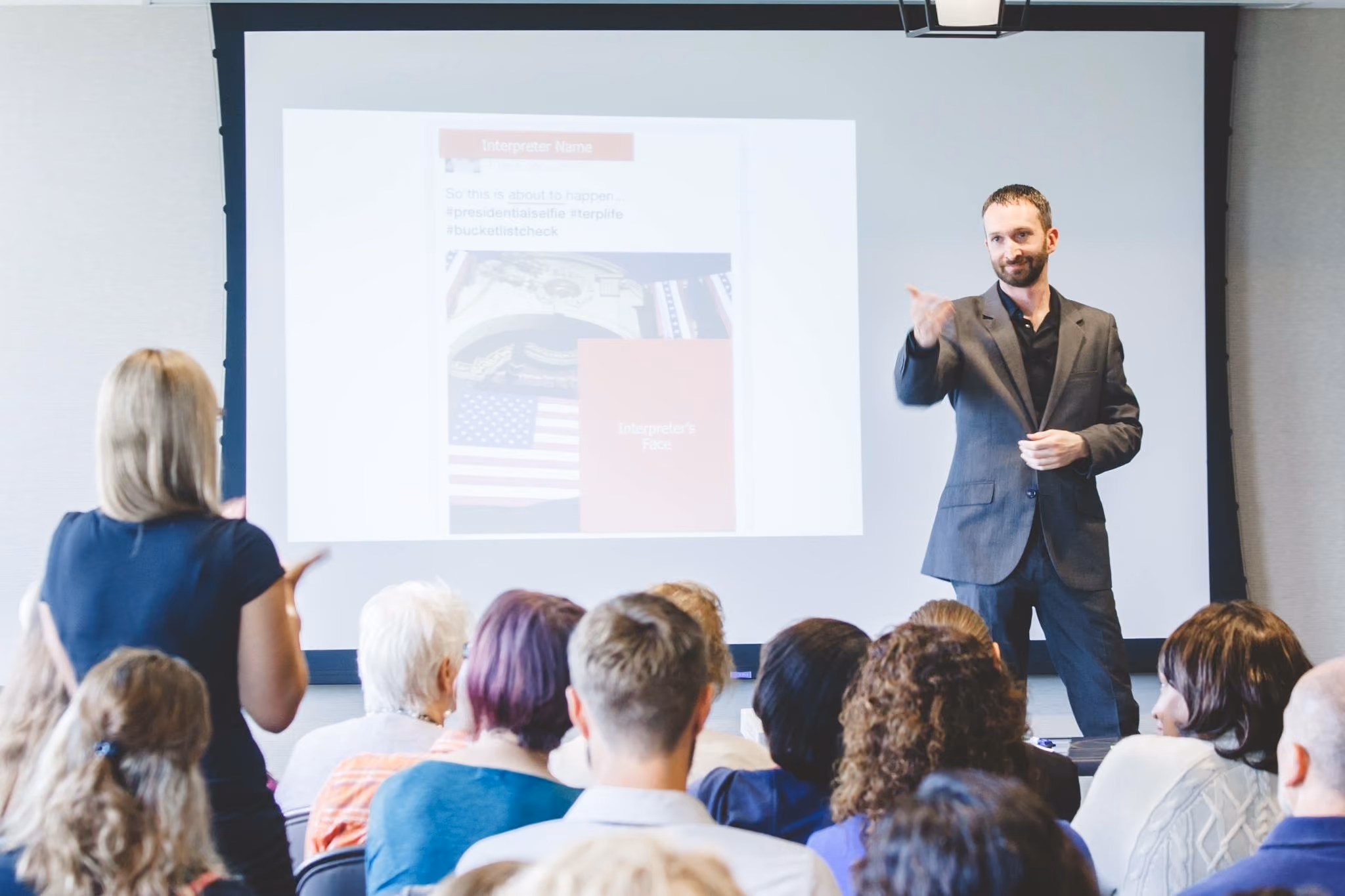 A man in a dark suit gives a presentation using American Sign Language (ASL) in front of an audience. A woman in the foreground signs toward him.