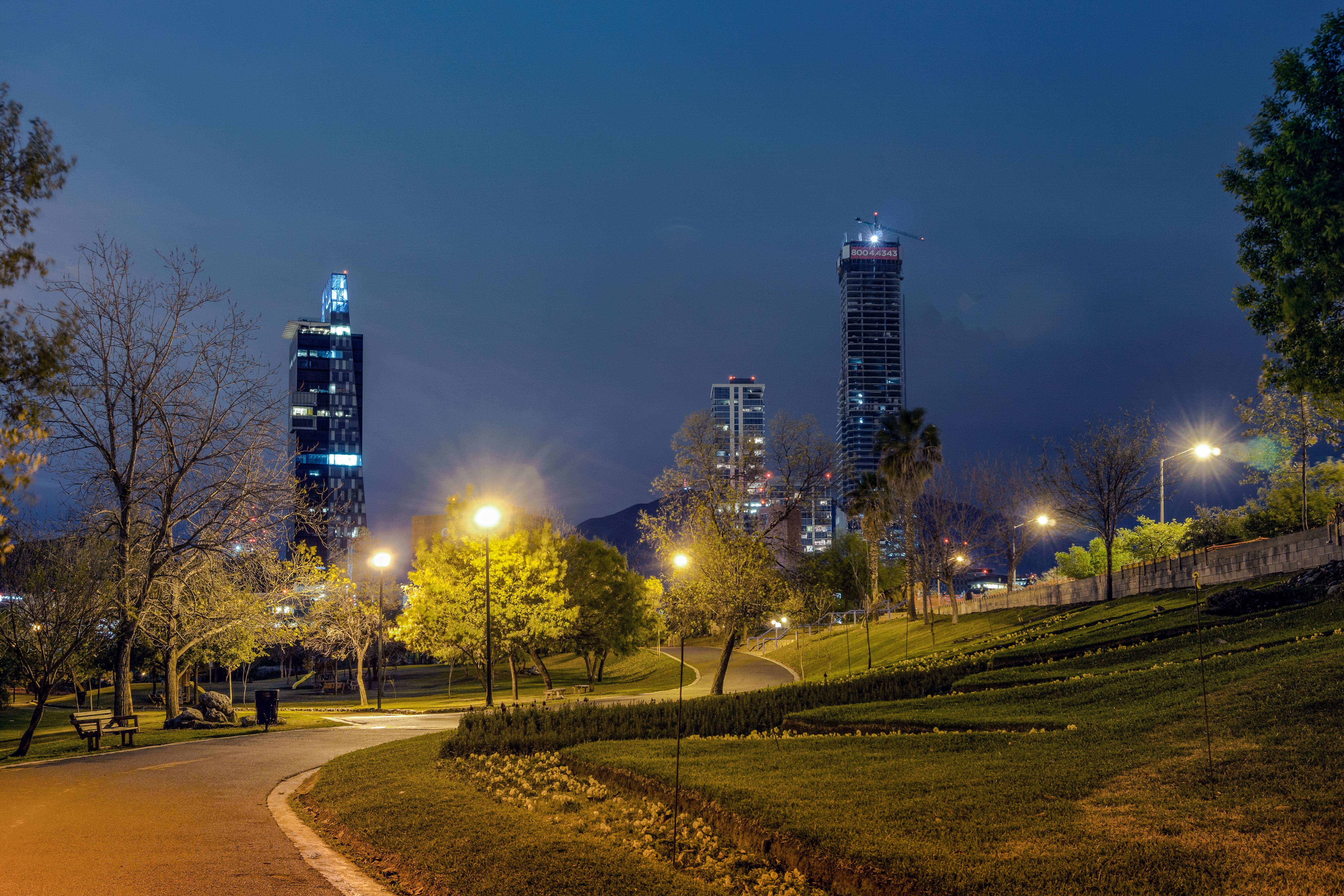 Camino iluminado y áreas verdes del Parque Rufino Tamayo, con edificios de Valle Oriente al fondo.