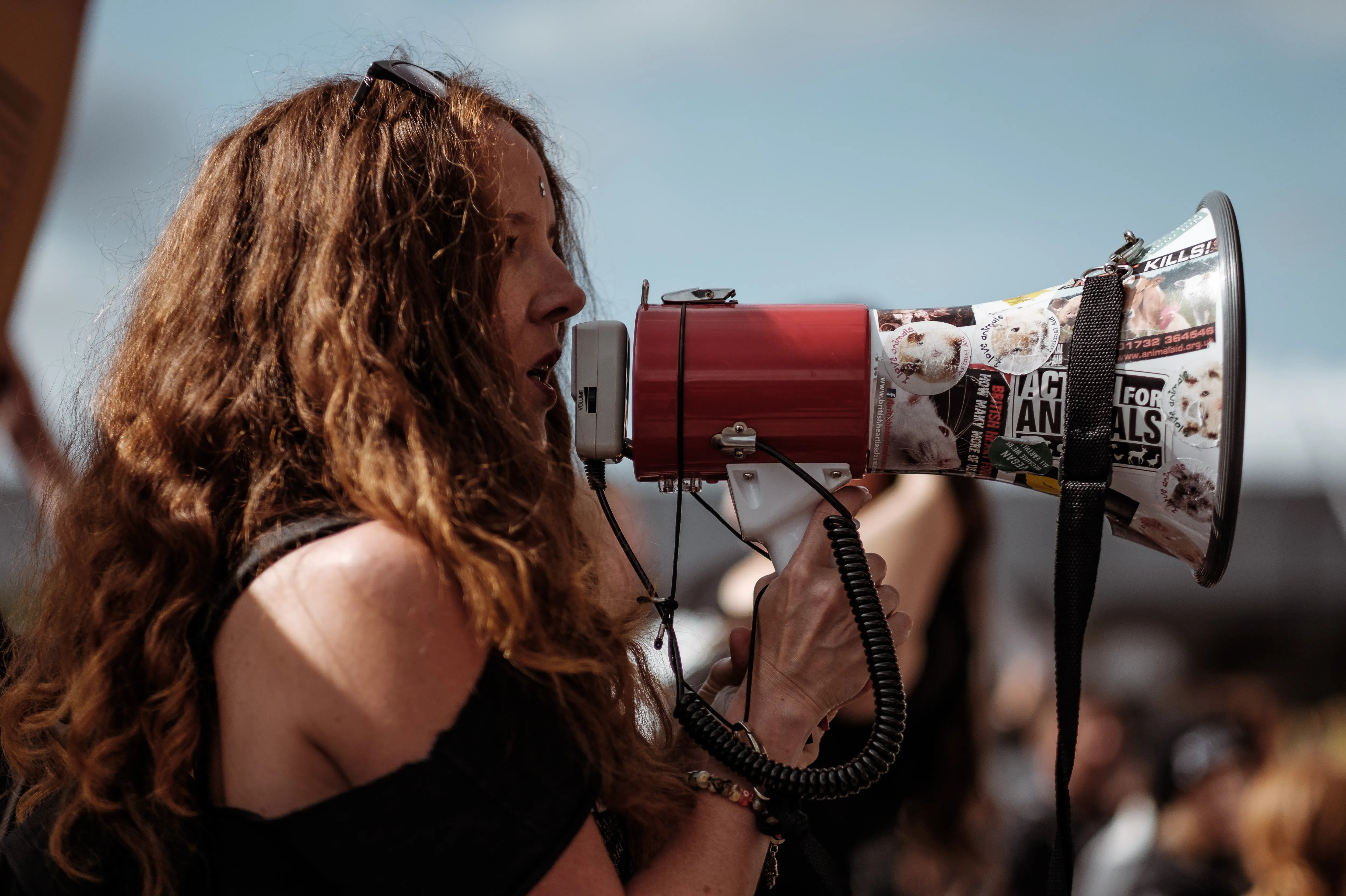 Woman with long hair passionately speaks into a red megaphone at a protest. The megaphone displays animal rights stickers. Blue sky in the background.