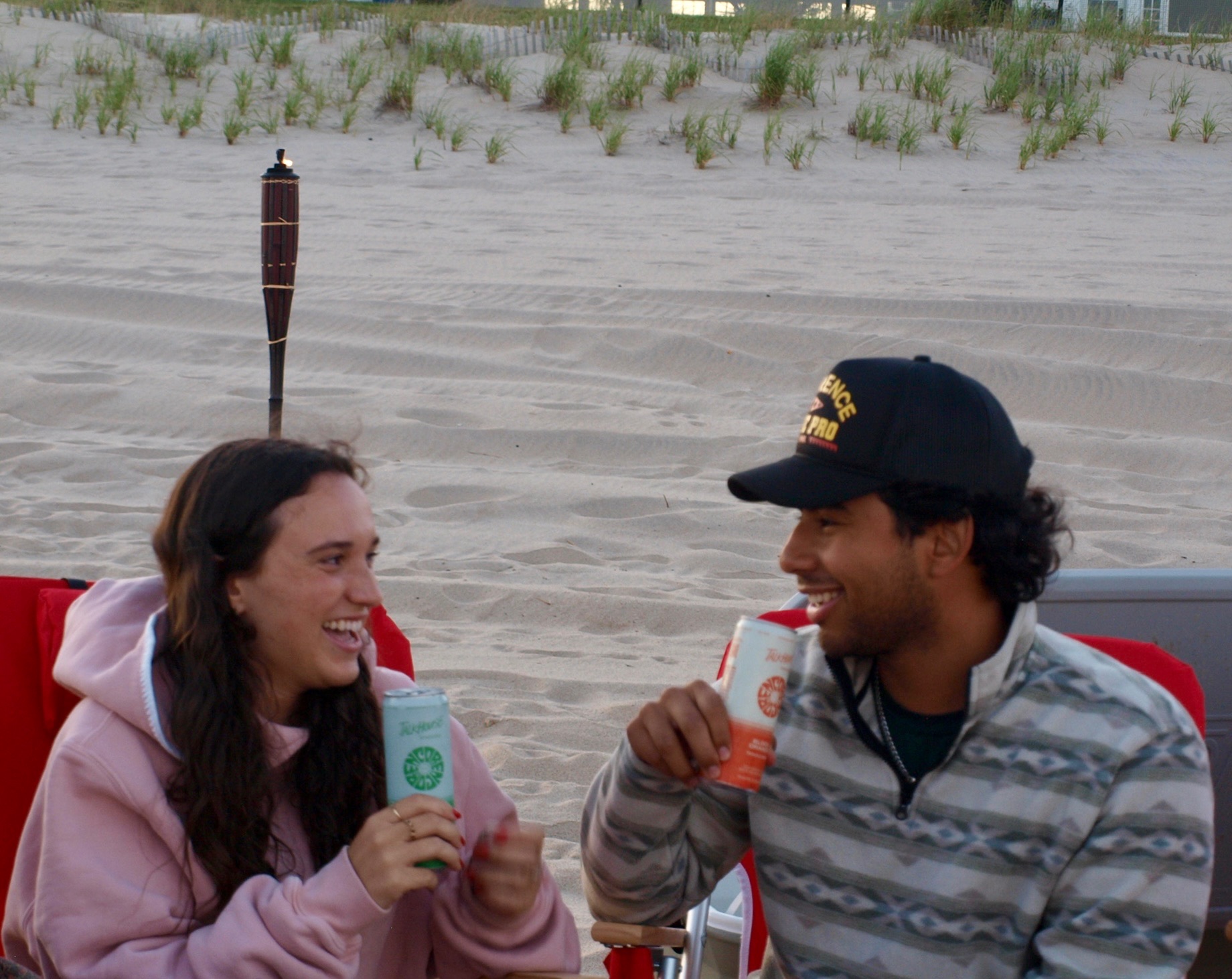 Two people laughing in beach chairs on the sand, each holding a can of Talkhouse Encore