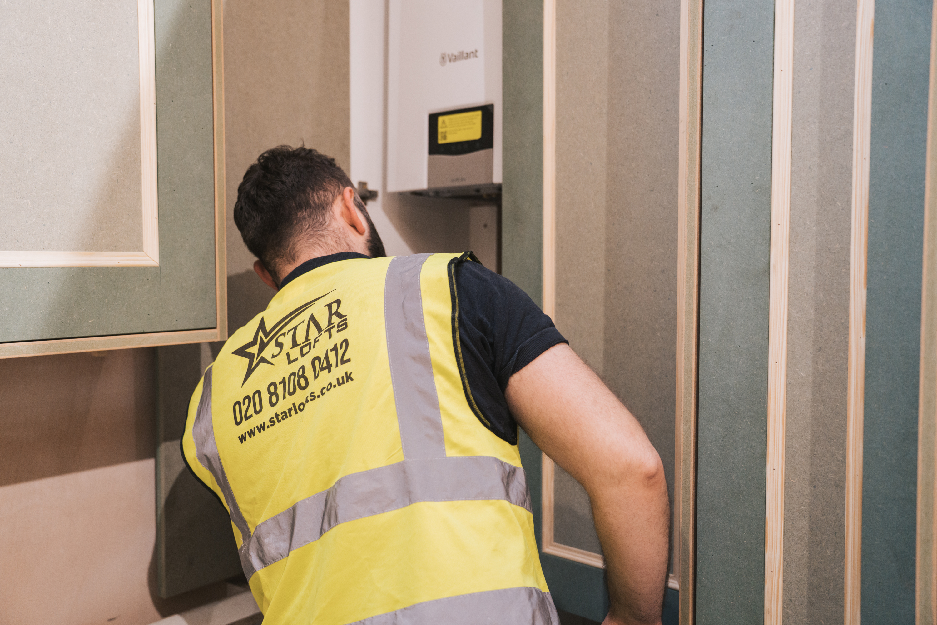 Worker in a yellow Star Lofts vest inspecting a wall-mounted boiler unit indoors.
