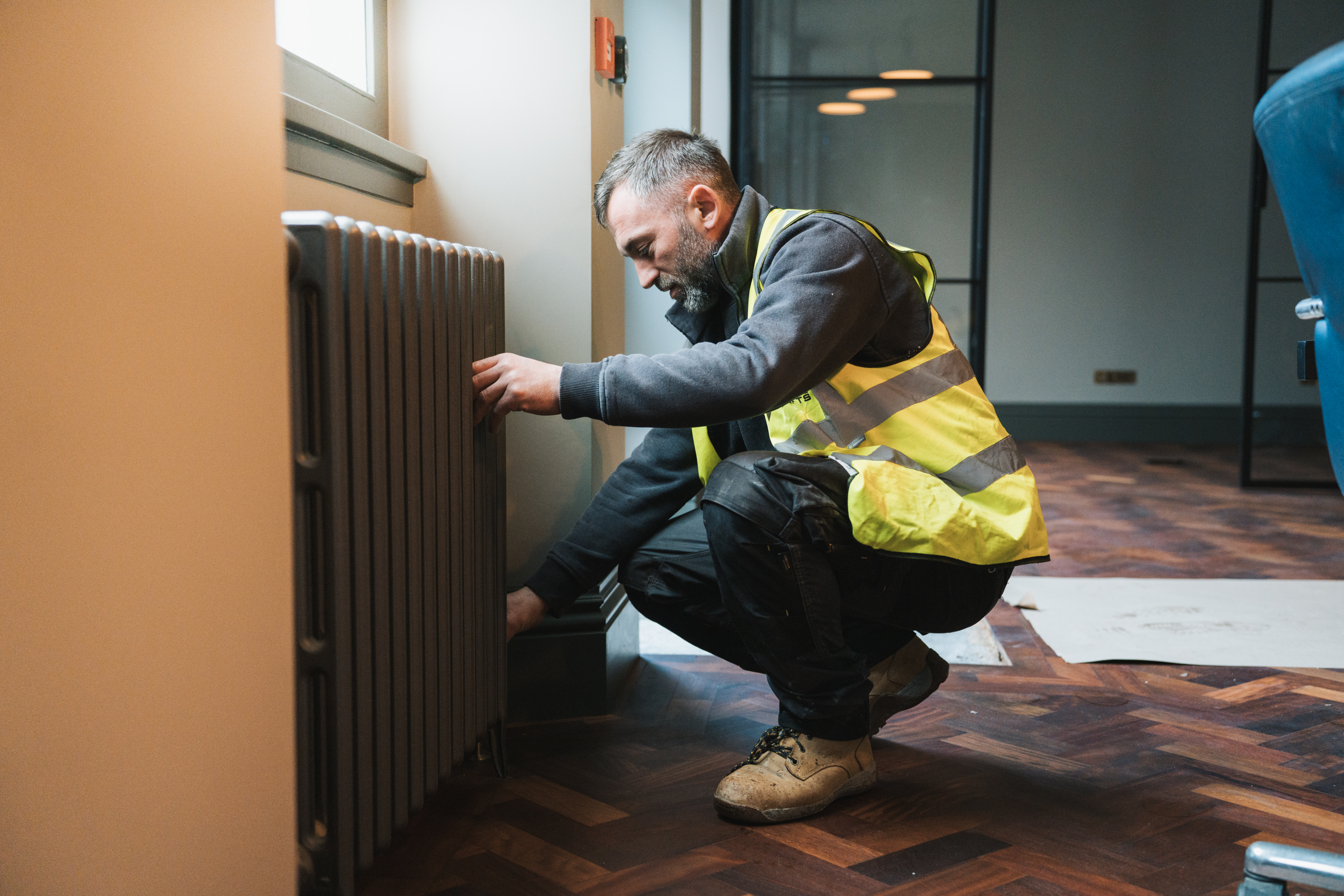 Worker in a yellow safety vest crouching and adjusting a radiator in a room with wooden floors.
