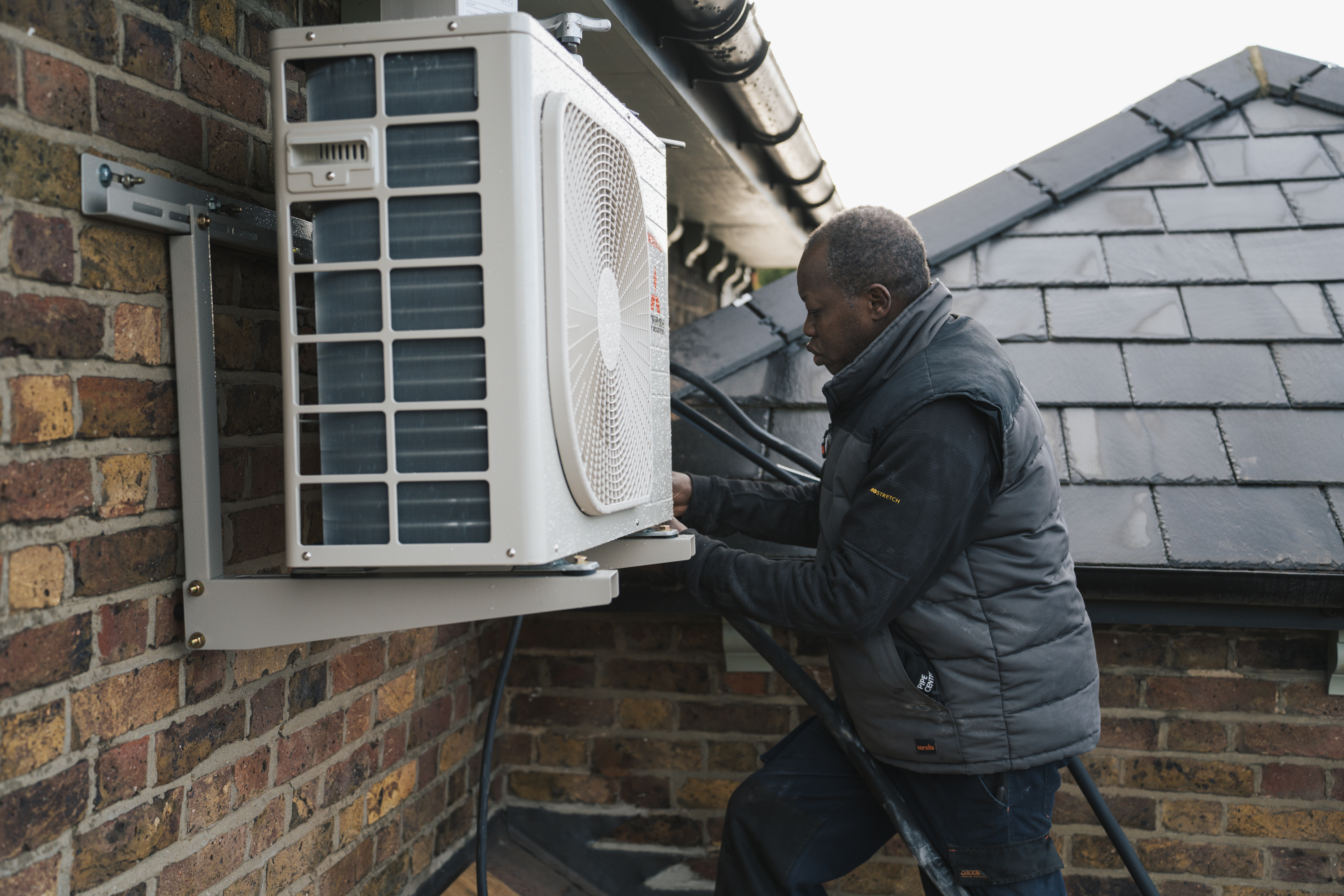 Man works on installing or repairing an outdoor air conditioning unit mounted on a brick wall.