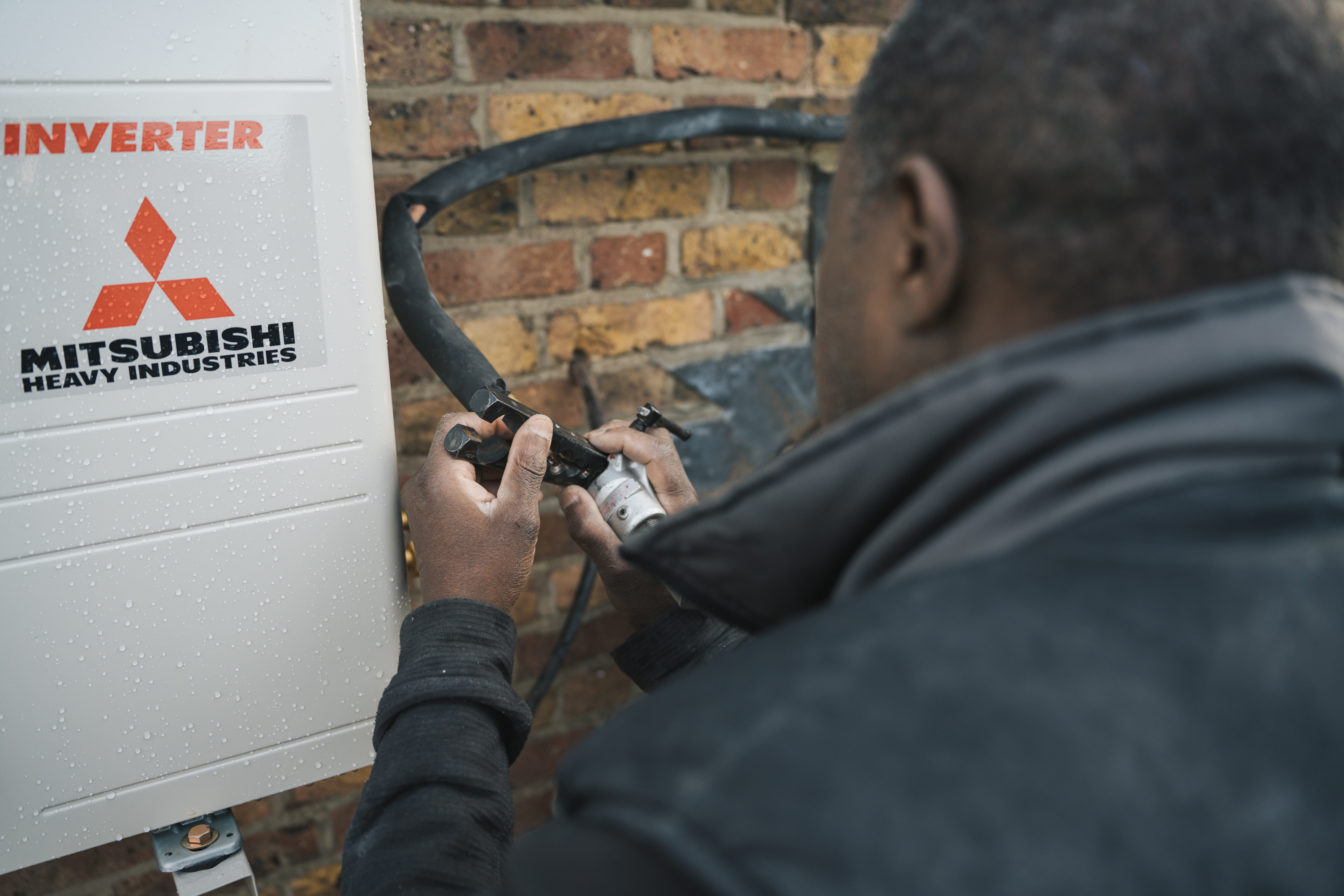 Technician installing or servicing a Mitsubishi Heavy Industries inverter on a brick wall outside.
