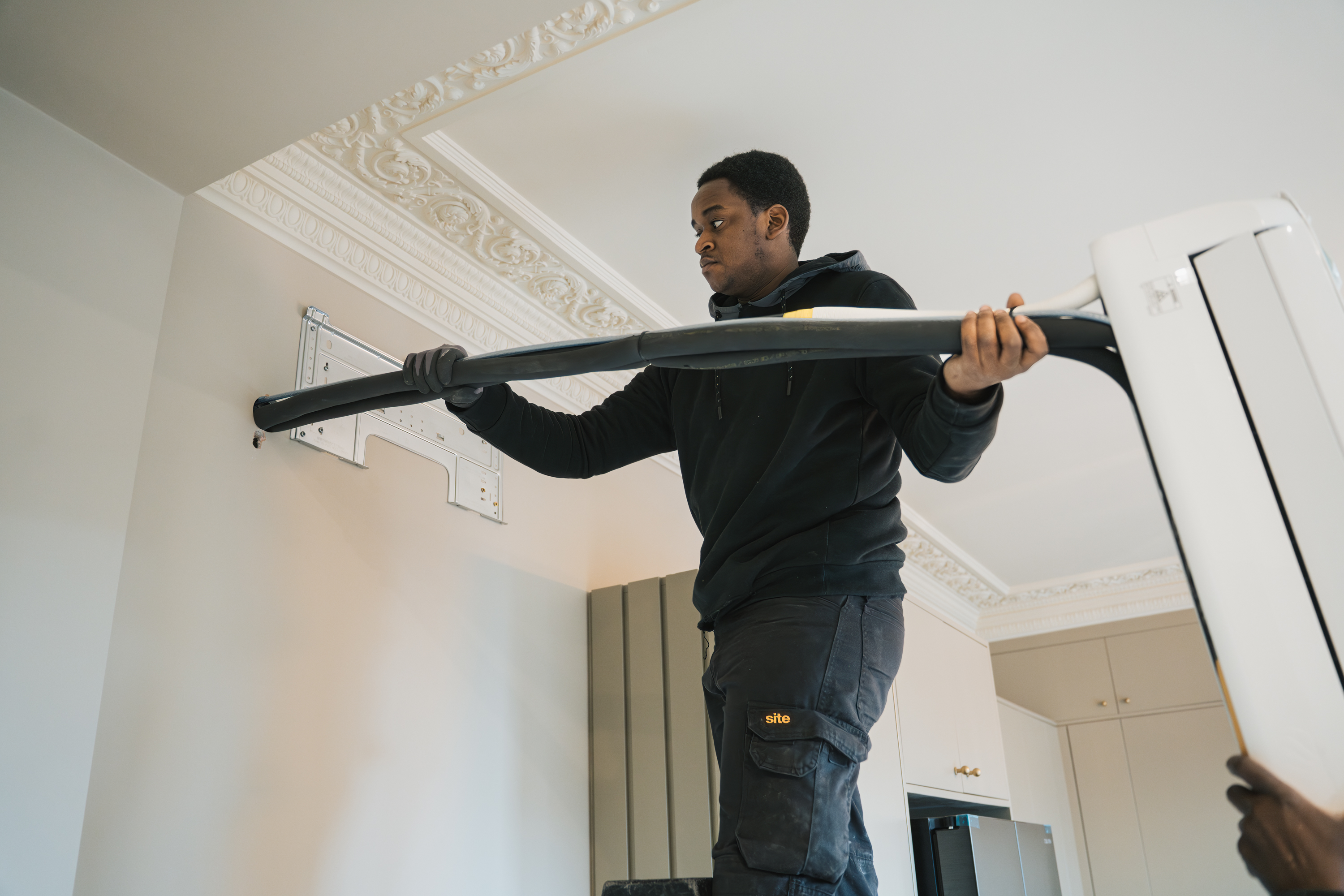 Technician in black installs a wall-mounted air conditioner inside a room with ornate ceiling trim.