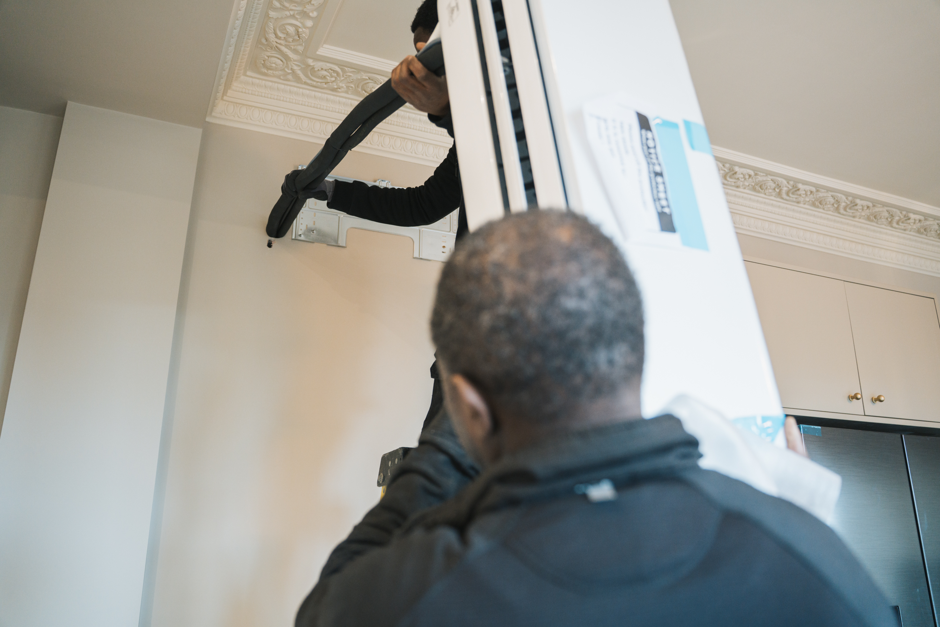Two men installing or mounting a large TV on a wall inside a room with decorated ceiling molding.